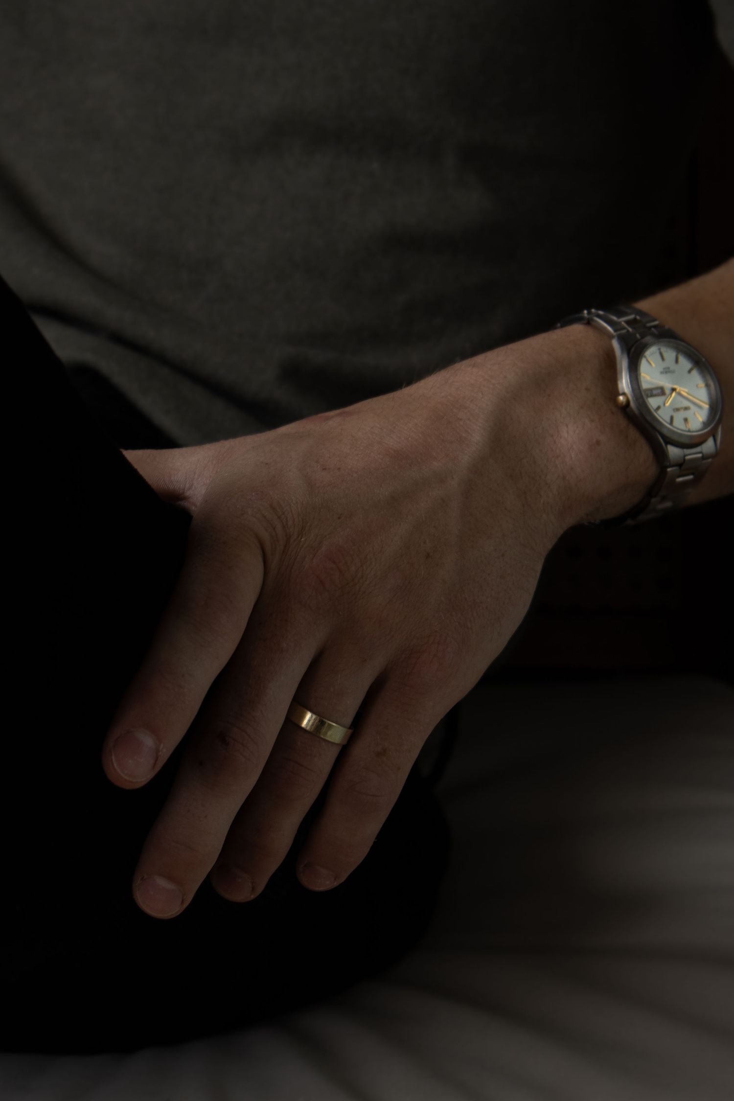 Close-up of a man's hand wearing a gold wedding band and silver watch, resting on his knee.