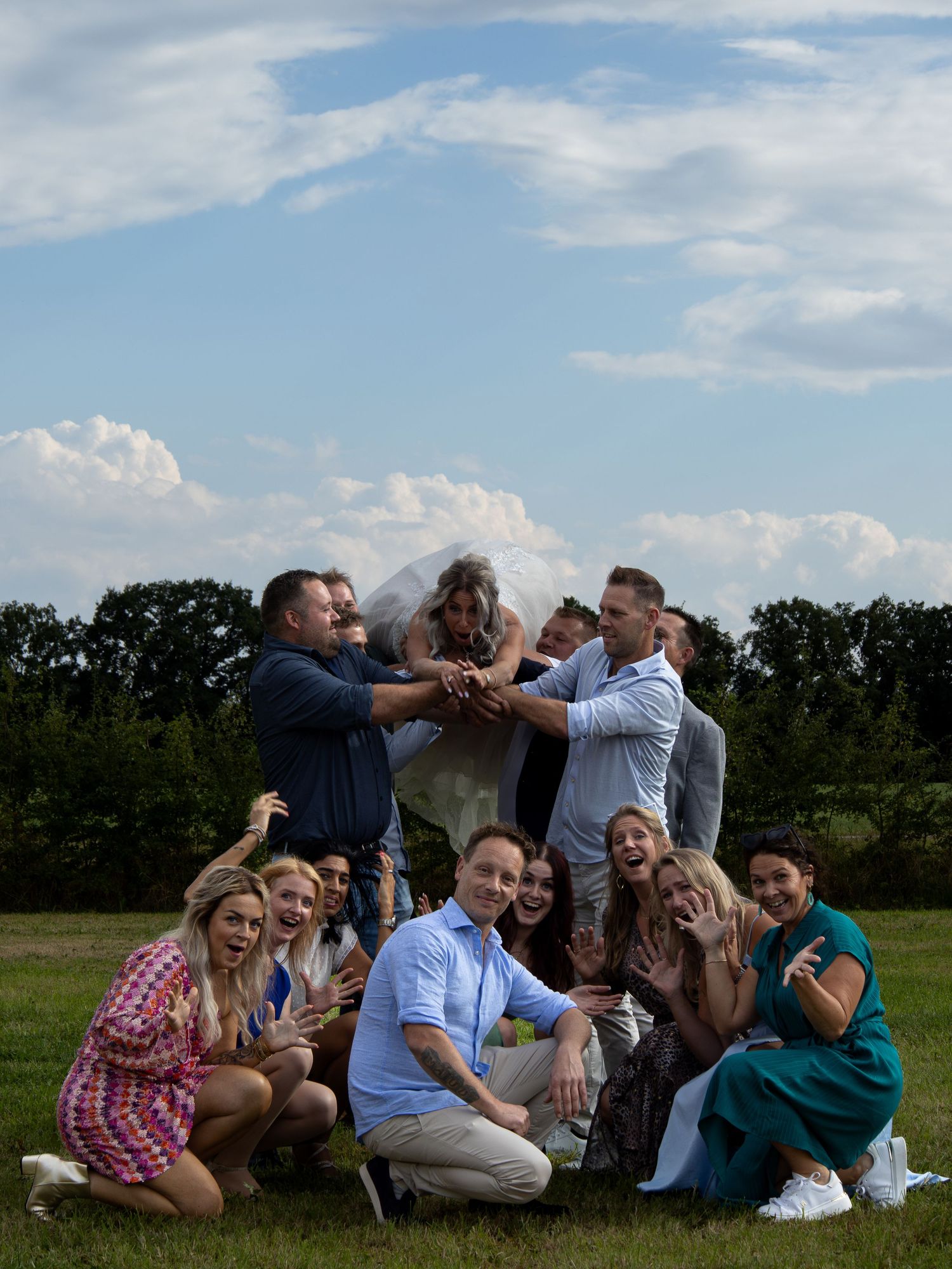 A joyful wedding party group photo outdoors, with bride and groom celebrated by friends on a green lawn under a blue sky.