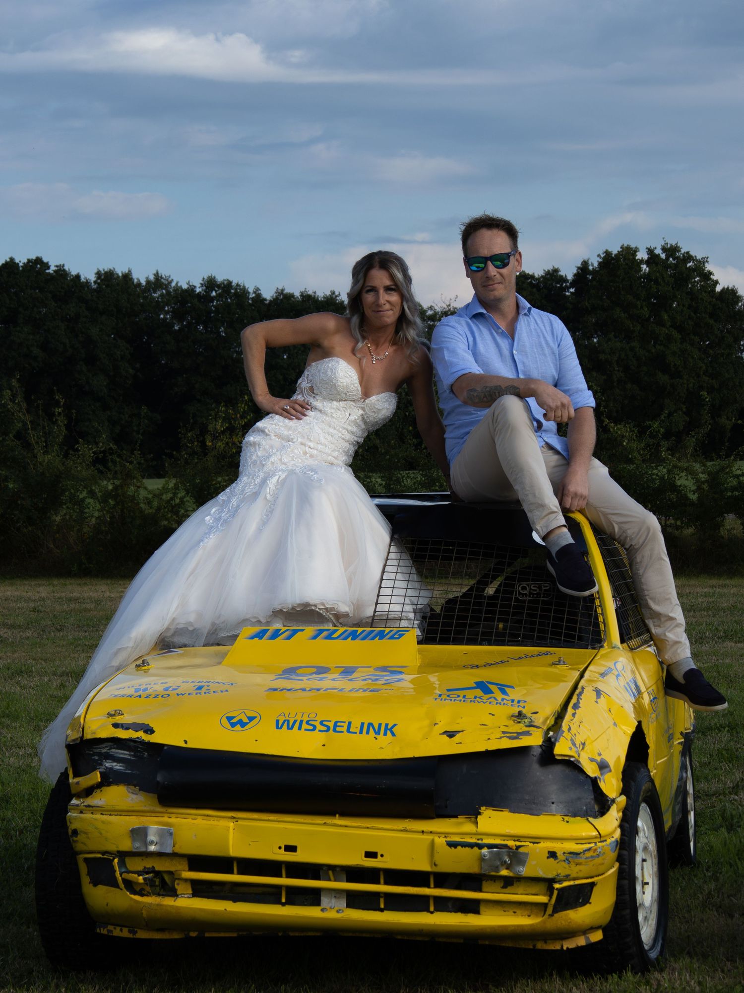 Bride in white gown and groom in blue shirt pose on a damaged yellow rally car outdoors.