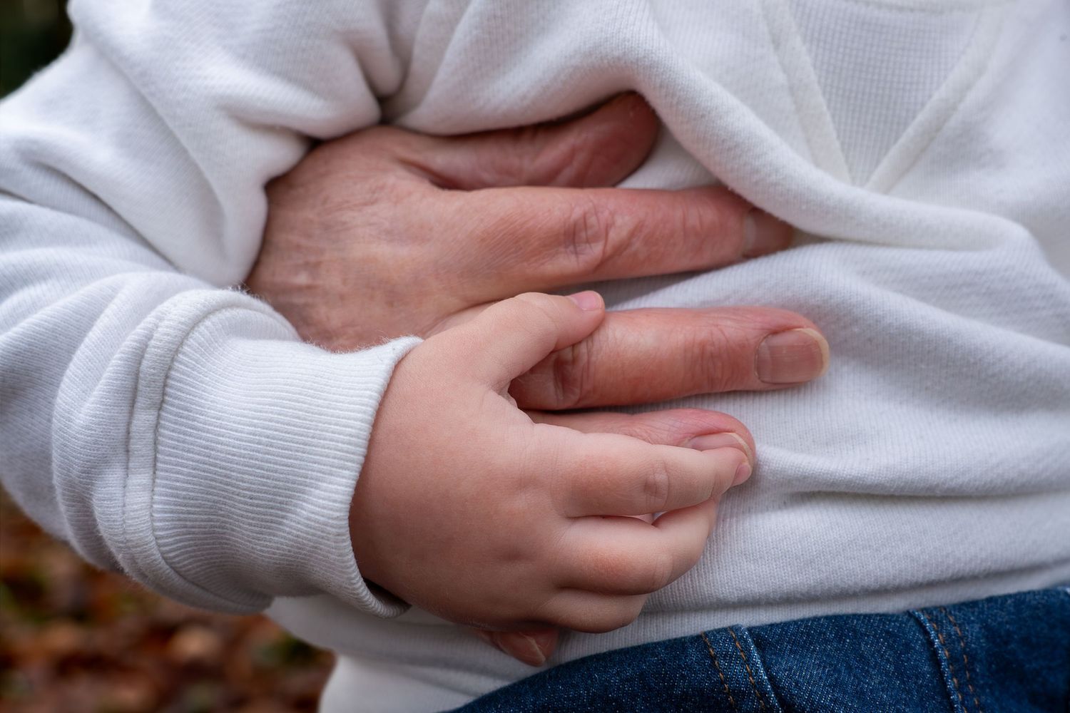Child clutching stomach in pain, wearing white hoodie and jeans, showing signs of abdominal discomfort outdoors.