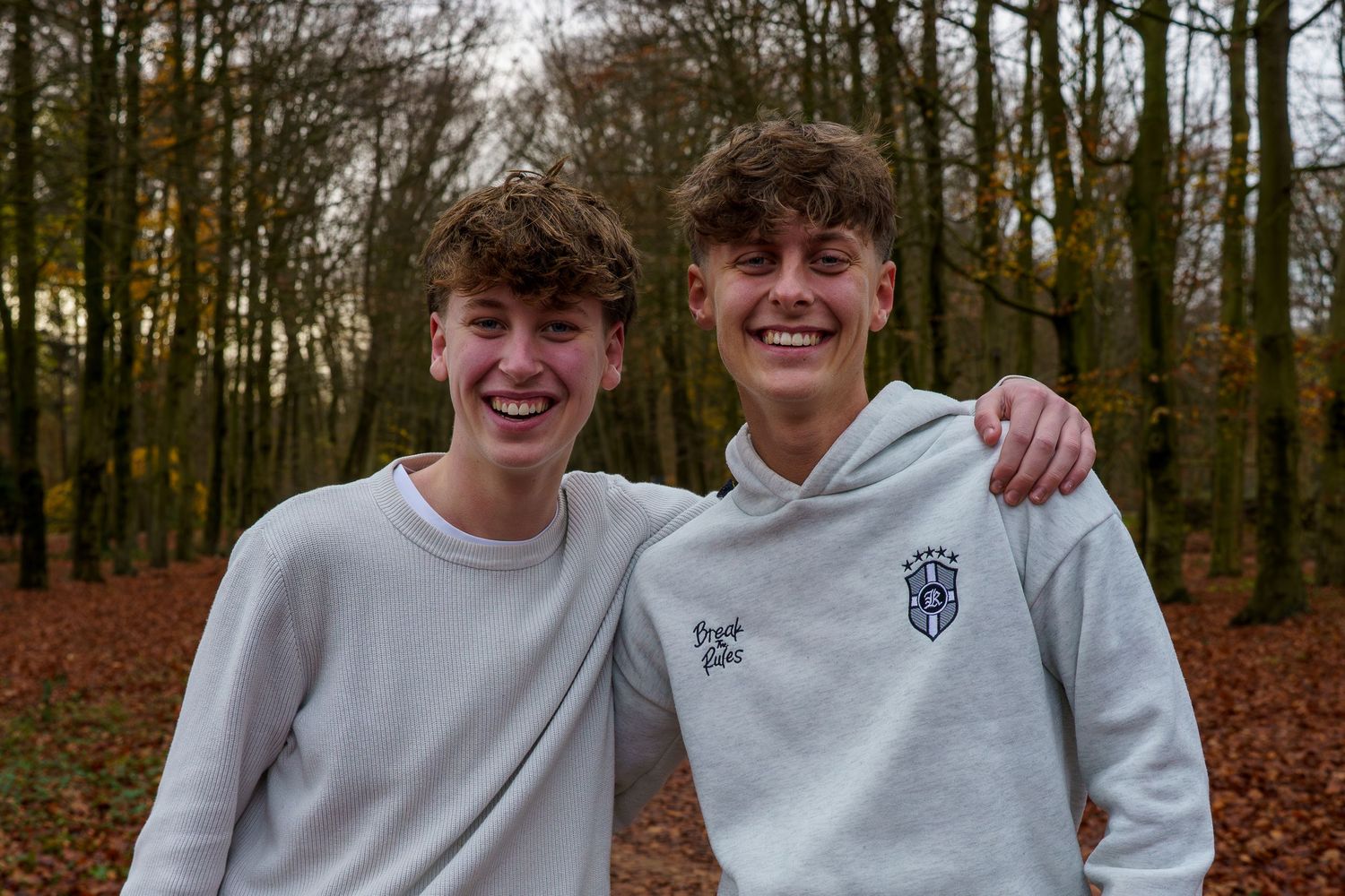 Two smiling teenage boys in grey hoodies pose together in an autumn forest with fallen leaves.
