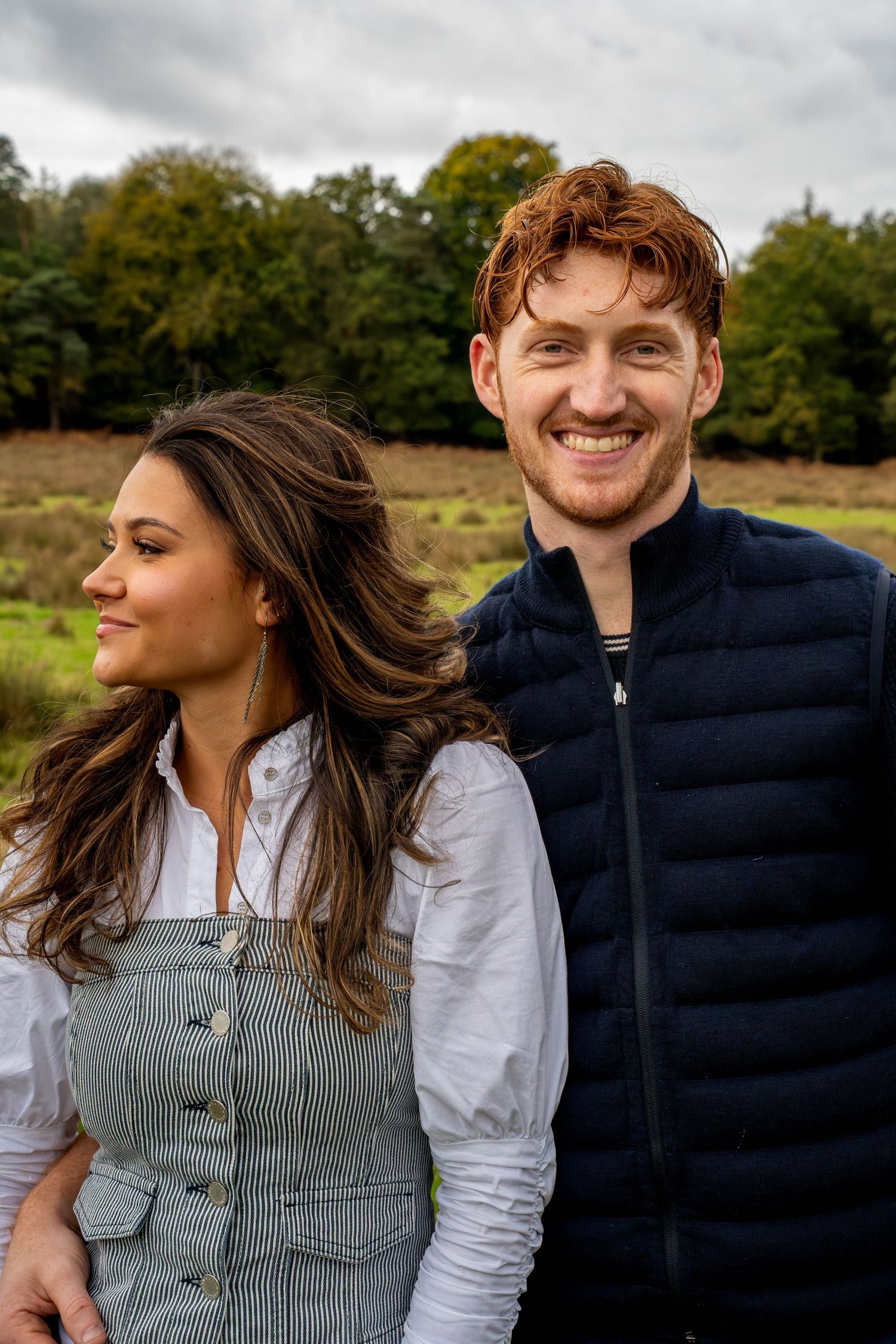 A smiling couple stands outdoors in a green field, the man in a navy vest and the woman in a denim pinafore.