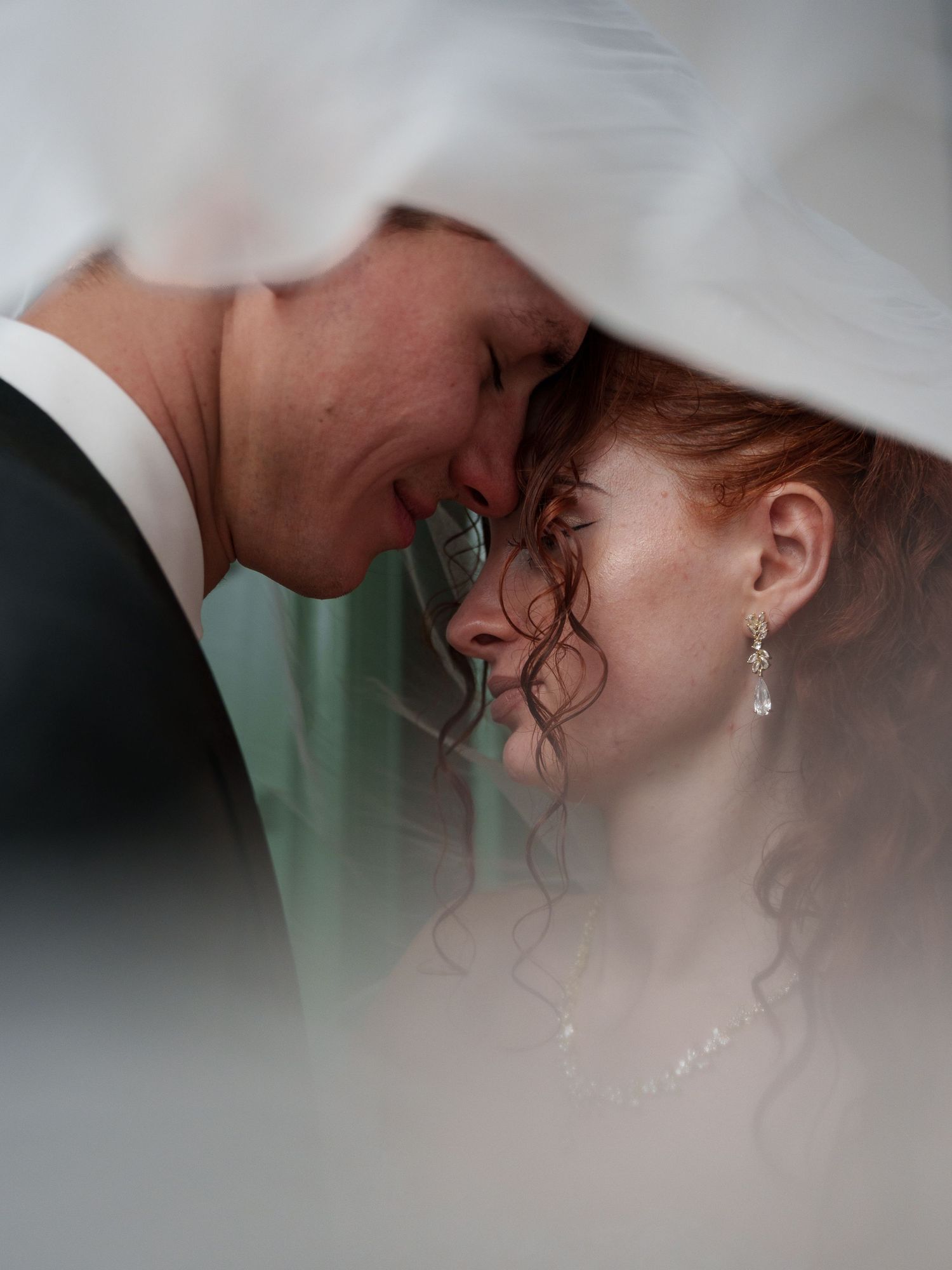 A bride and groom share an intimate forehead-to-forehead moment under a flowing white veil on their wedding day.