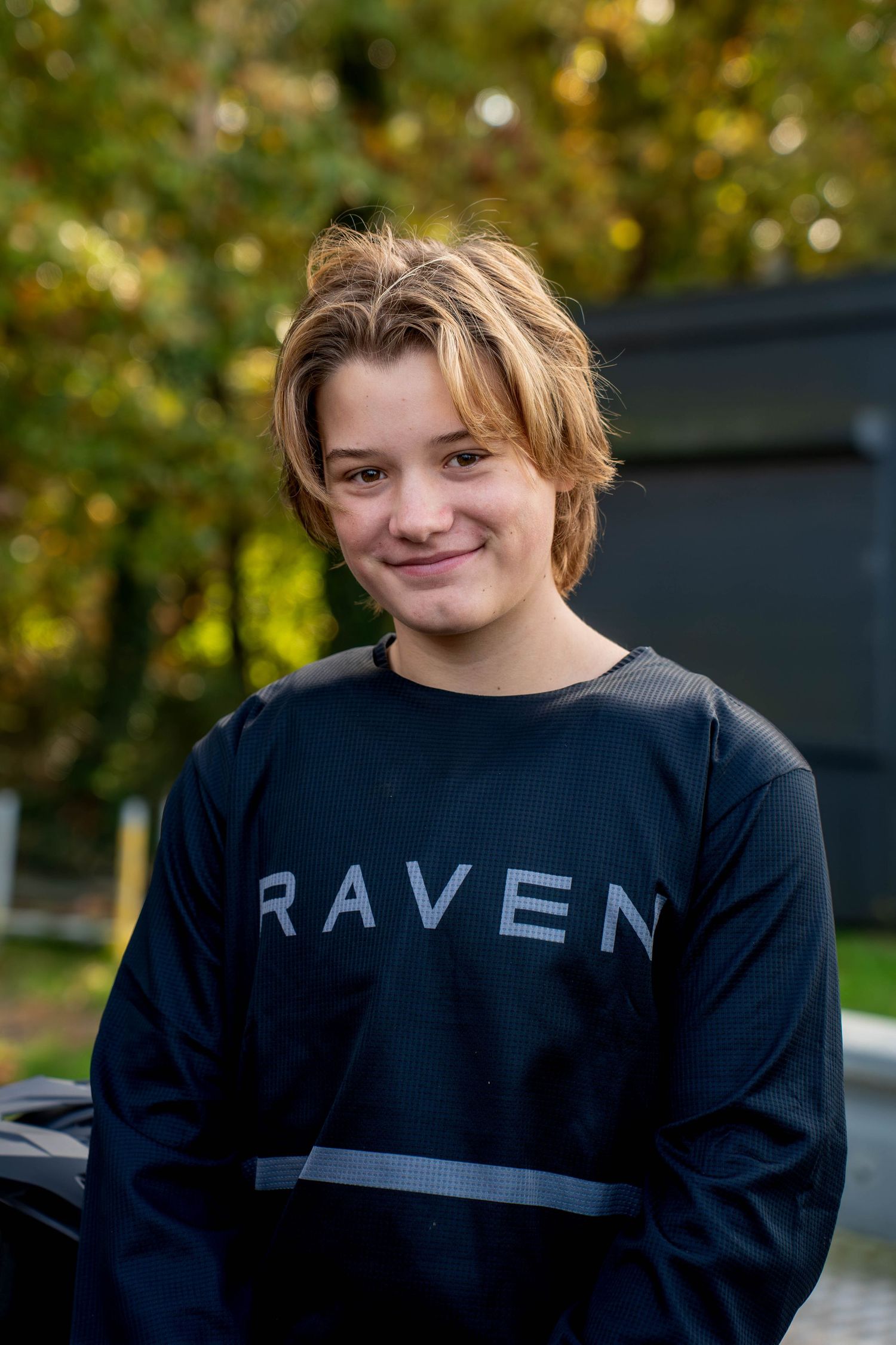 A person wearing a black Raven sweatshirt poses outdoors on a sunny autumn day with trees in the background.