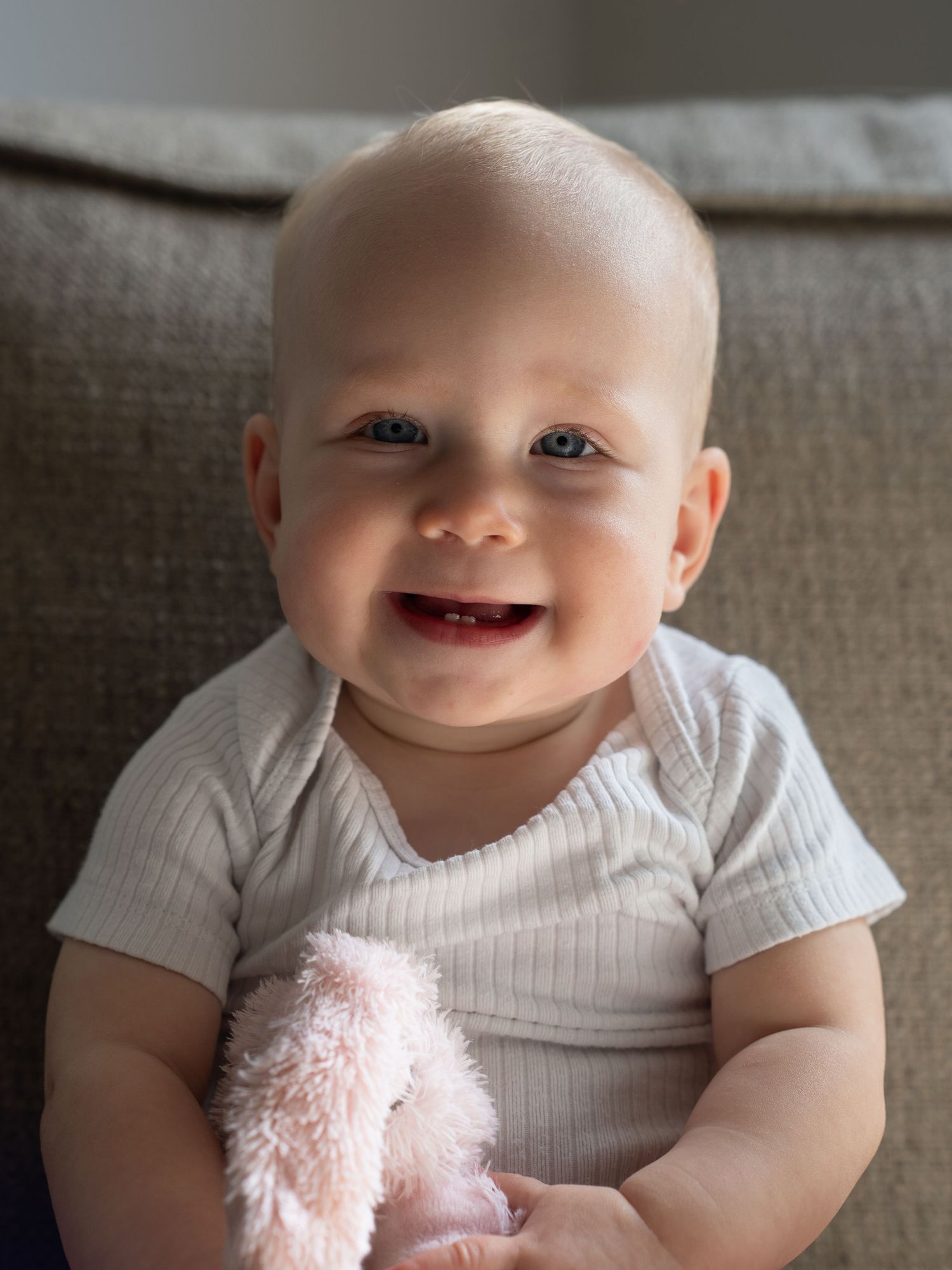 A baby wearing a white shirt smiles brightly while holding a pink plush toy.
