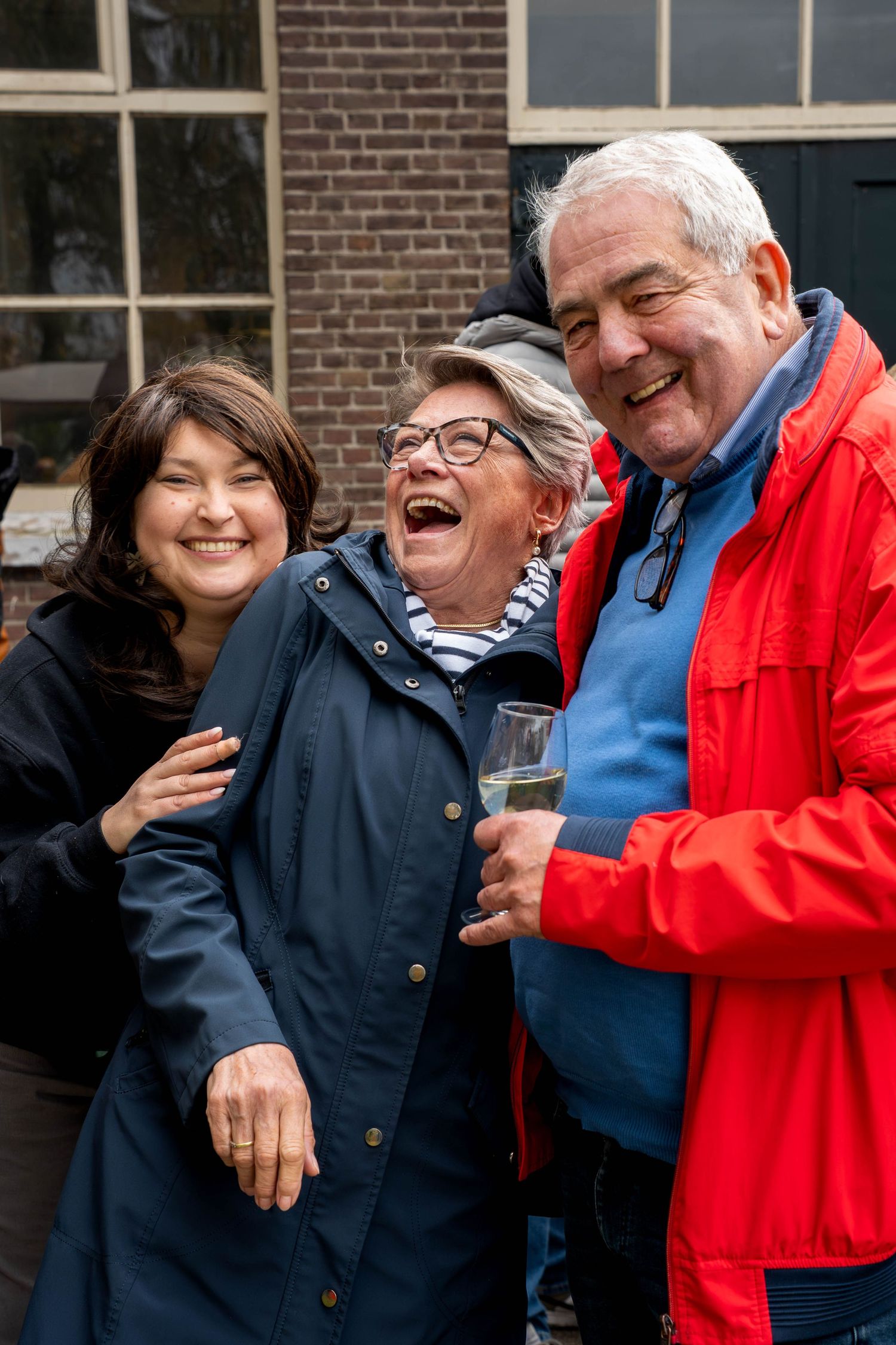 Three people laughing outdoors, older woman in navy jacket, man in red coat holding wine glass, younger woman smiling.