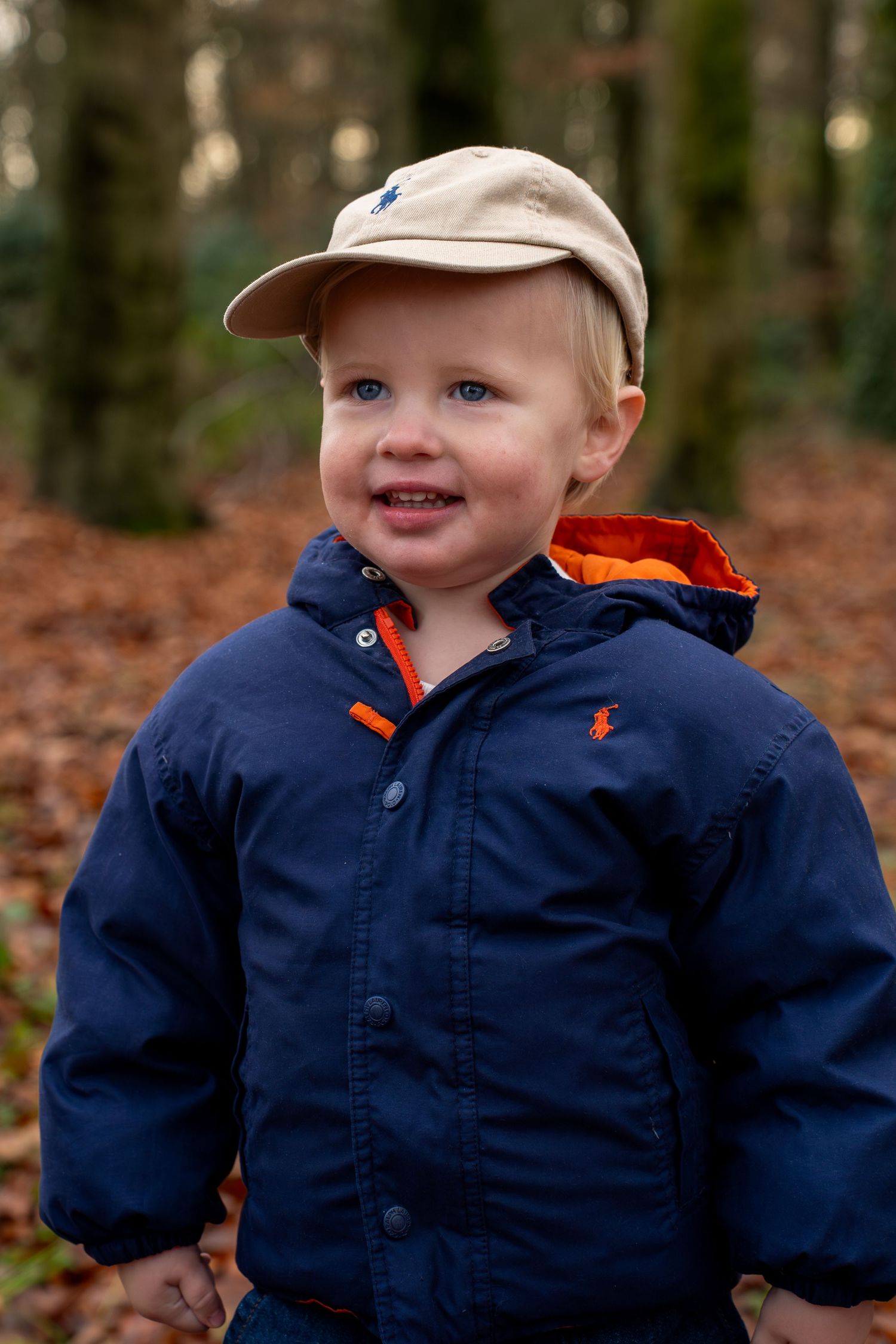 Toddler boy in navy Ralph Lauren jacket and beige cap smiling in autumn forest with fallen leaves.