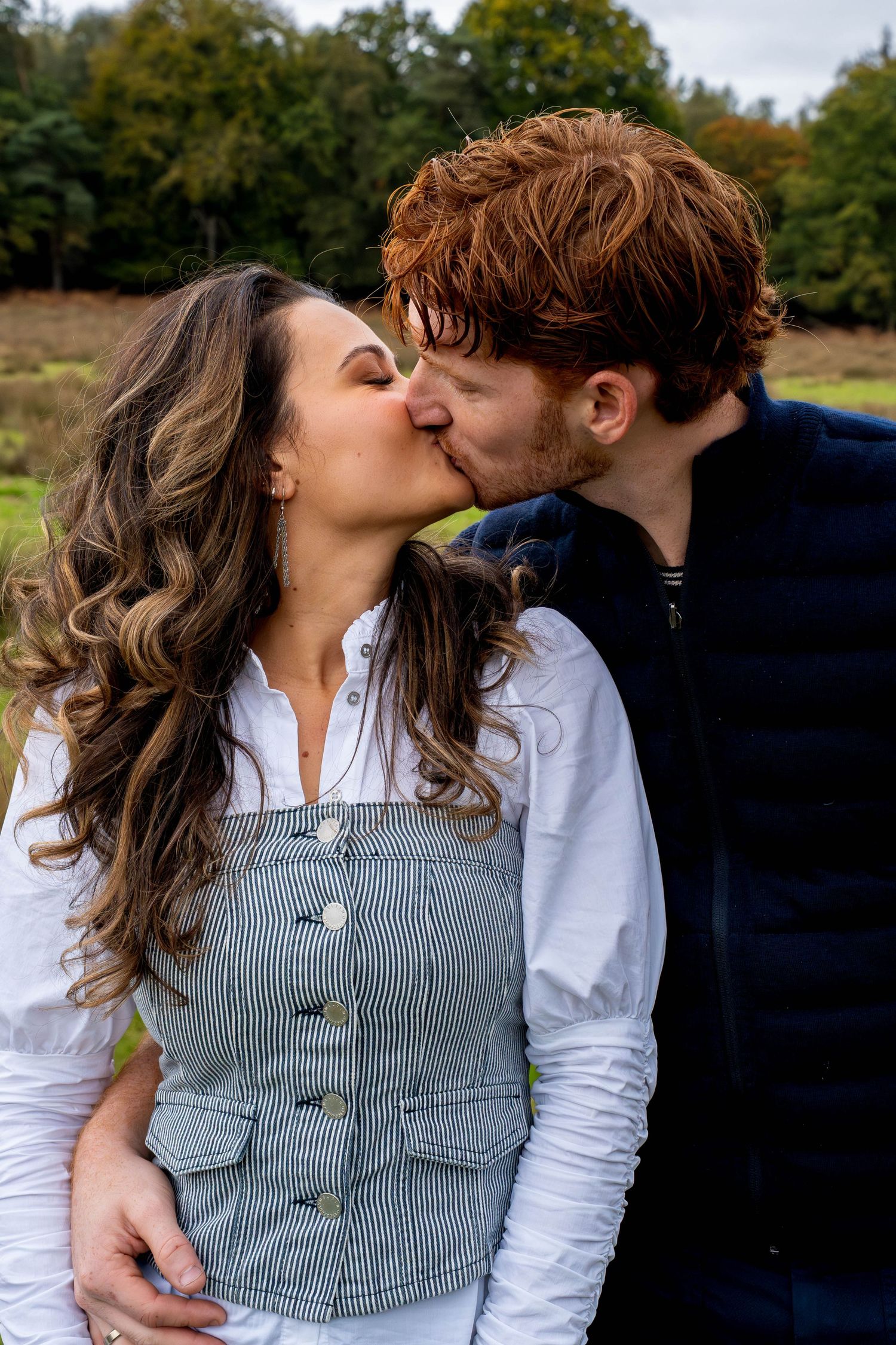 A couple sharing a romantic kiss outdoors, surrounded by autumn trees in a lush green field.
