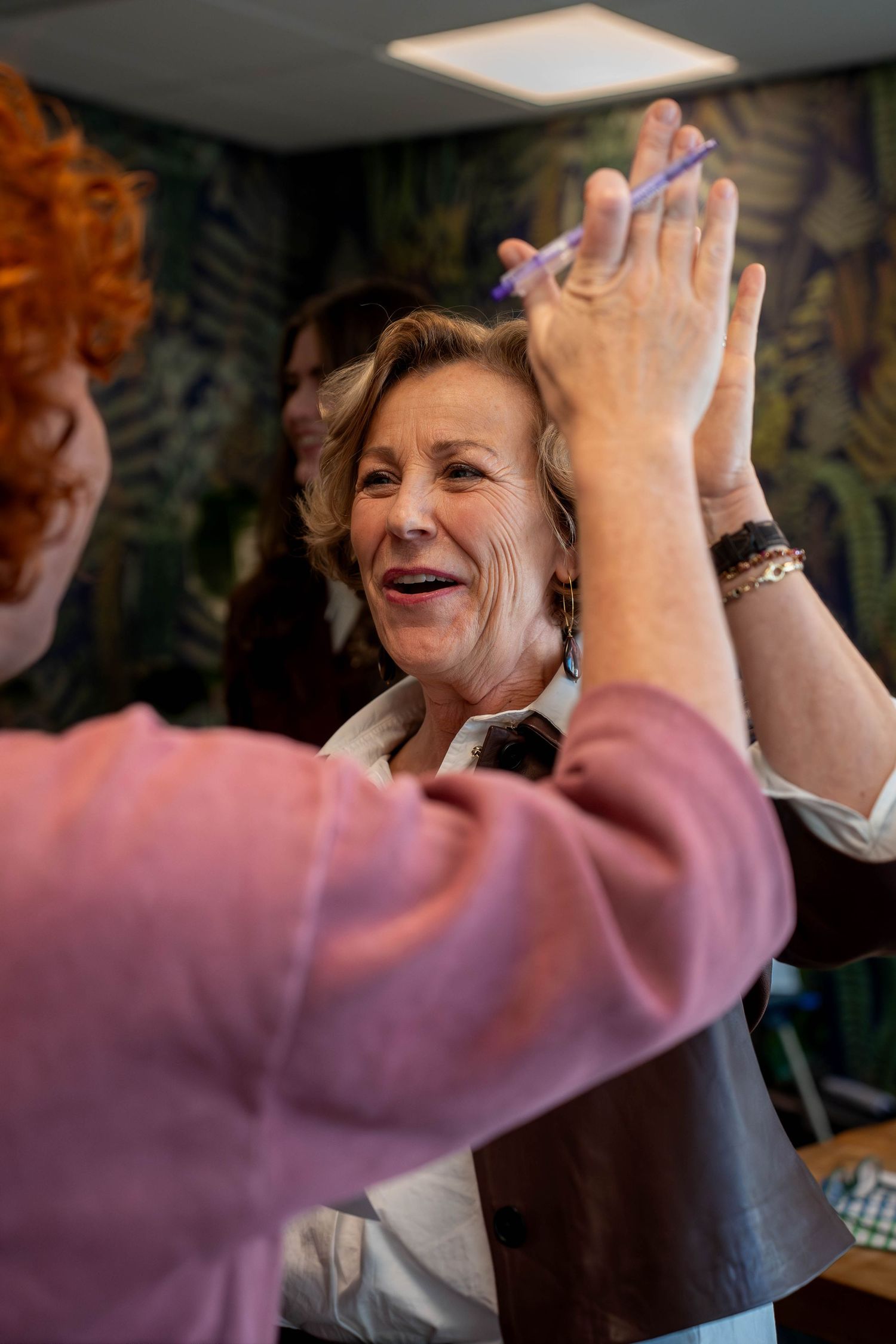 A hairstylist with red hair cuts the hair of a smiling older woman in a salon with green floral wallpaper.