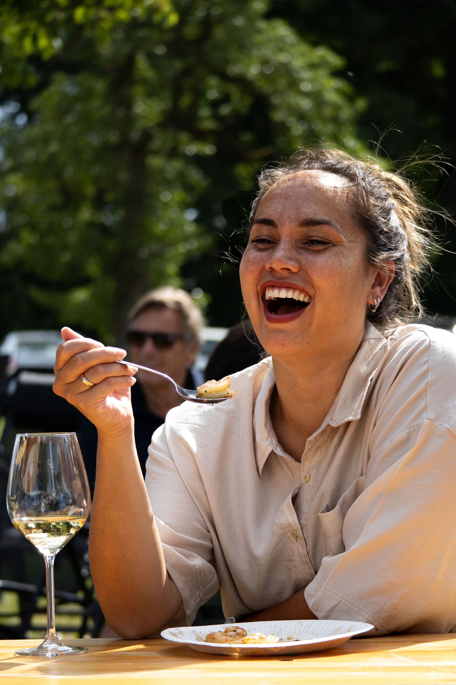 A person laughs joyfully while sitting at an outdoor table with a glass of white wine on a sunny day.