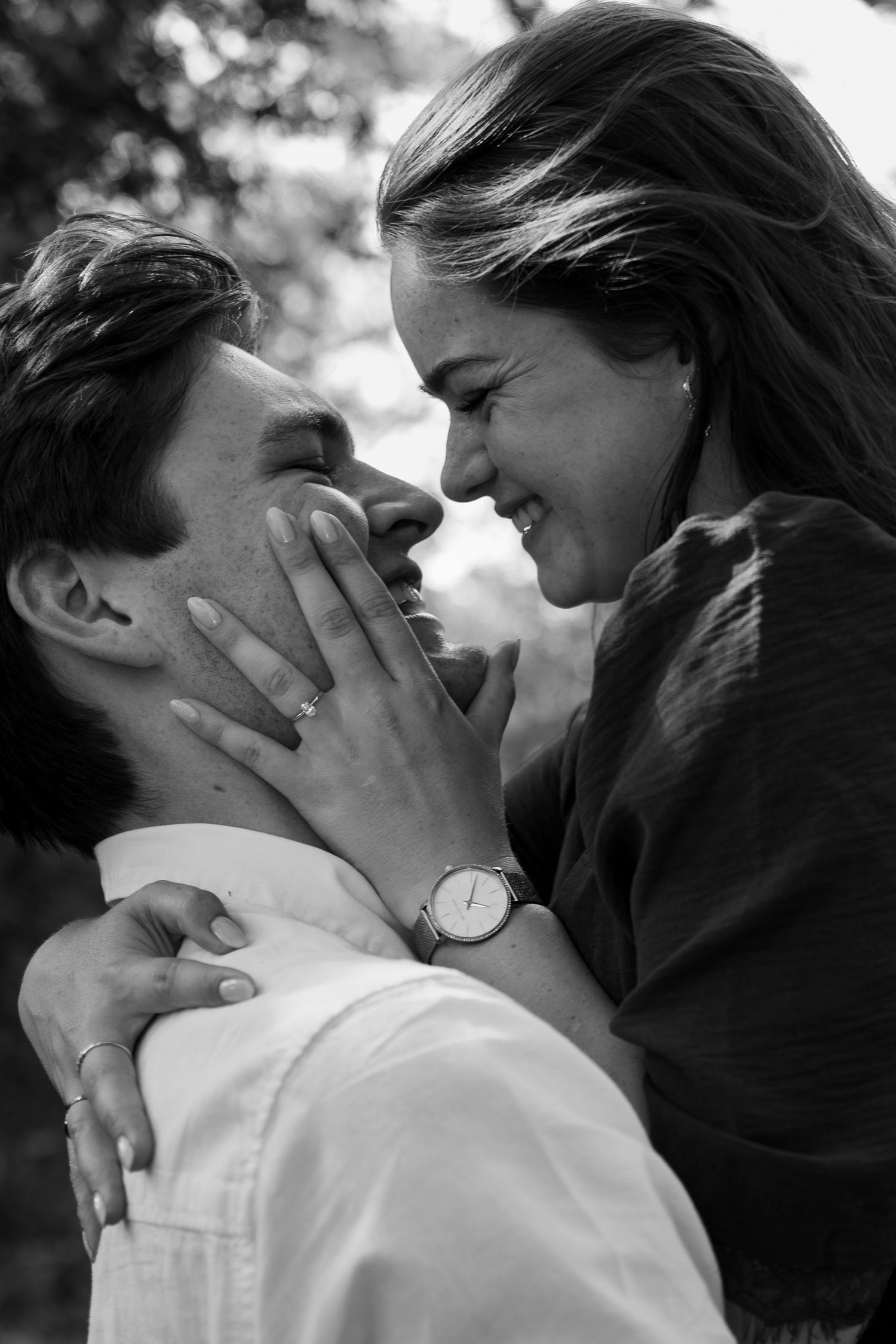 Black and white photo of a couple smiling and embracing outdoors, woman wearing a watch and ring touching mans face.