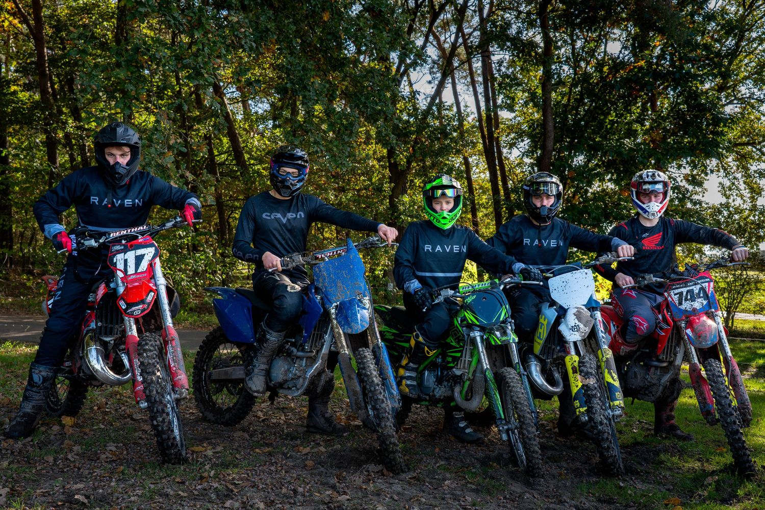 Five dirt bike riders in helmets and gear pose with their motorcycles in a wooded outdoor setting.