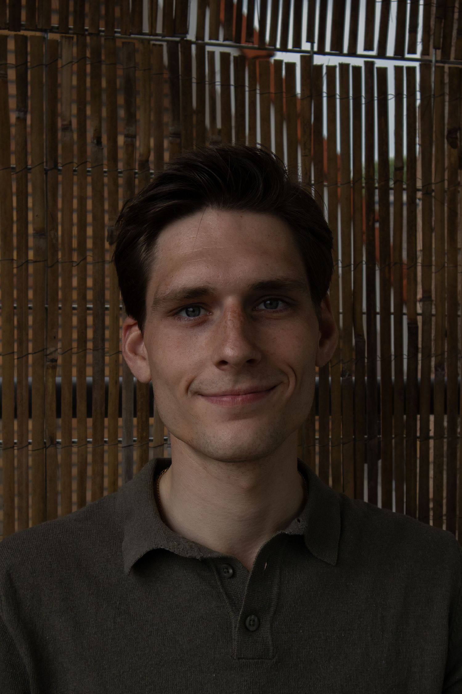 Young man with blue eyes in olive polo shirt smiling against a wooden slat wall background.