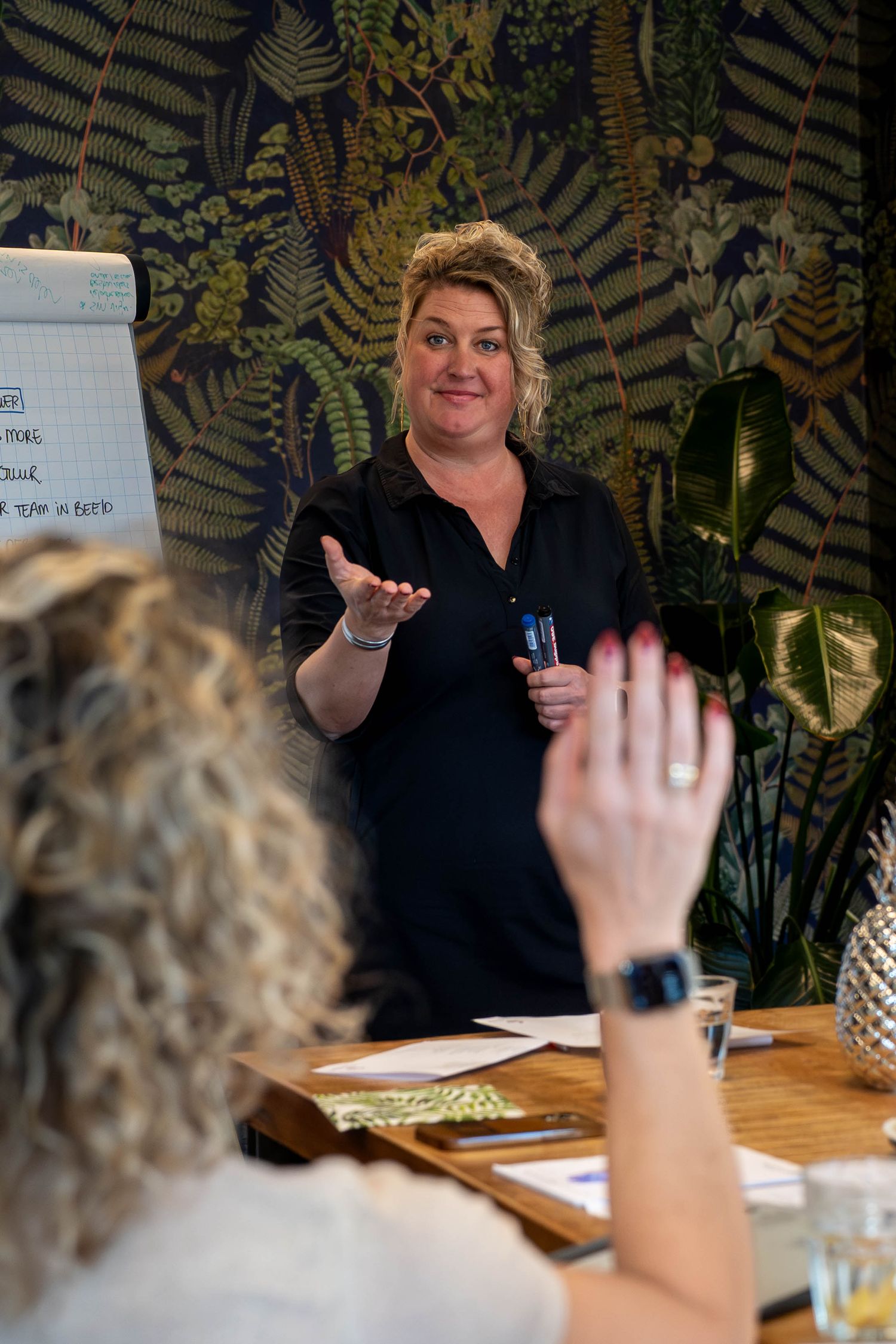 Woman presenting at business meeting with botanical wallpaper while participant raises hand.