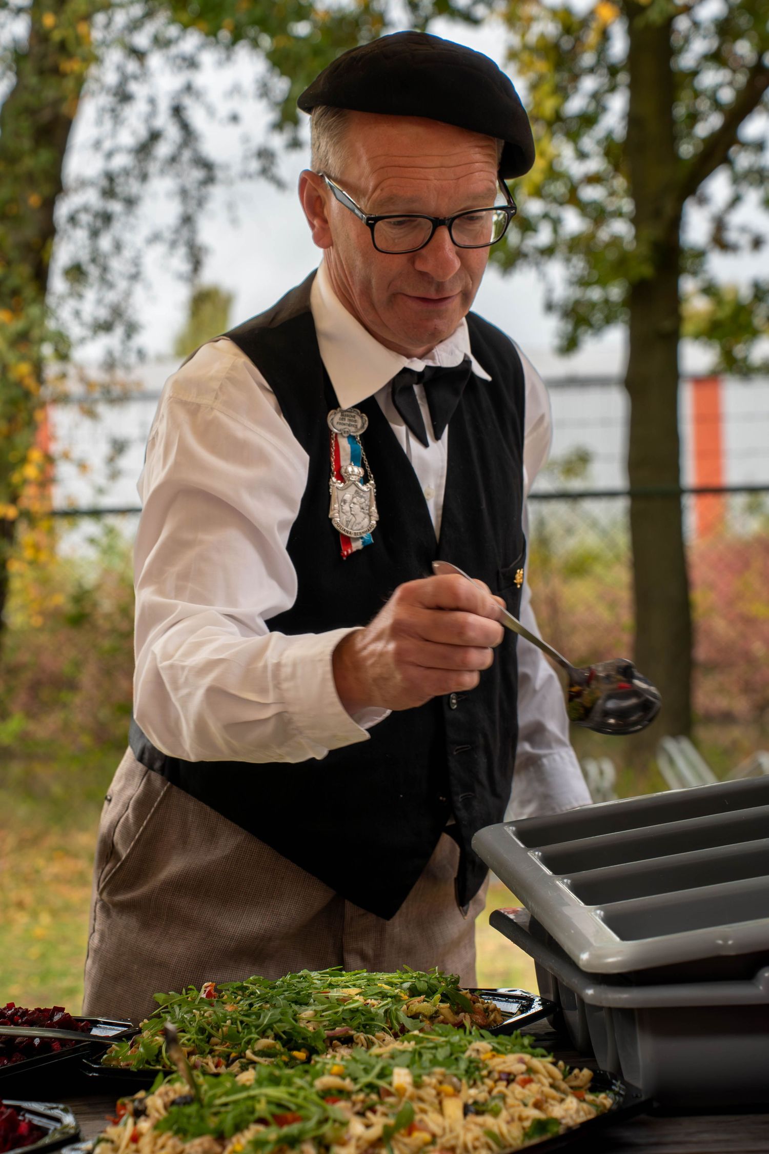A person in a black vest and white shirt examines food in a serving tray outdoors during autumn.
