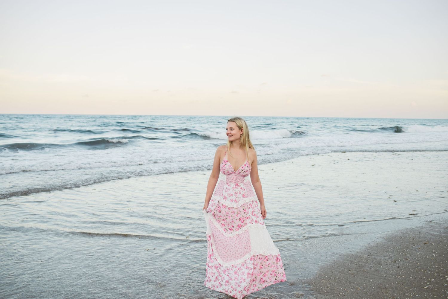 Blonde woman in a flowing pink floral maxi dress standing at the ocean's edge at sunset.