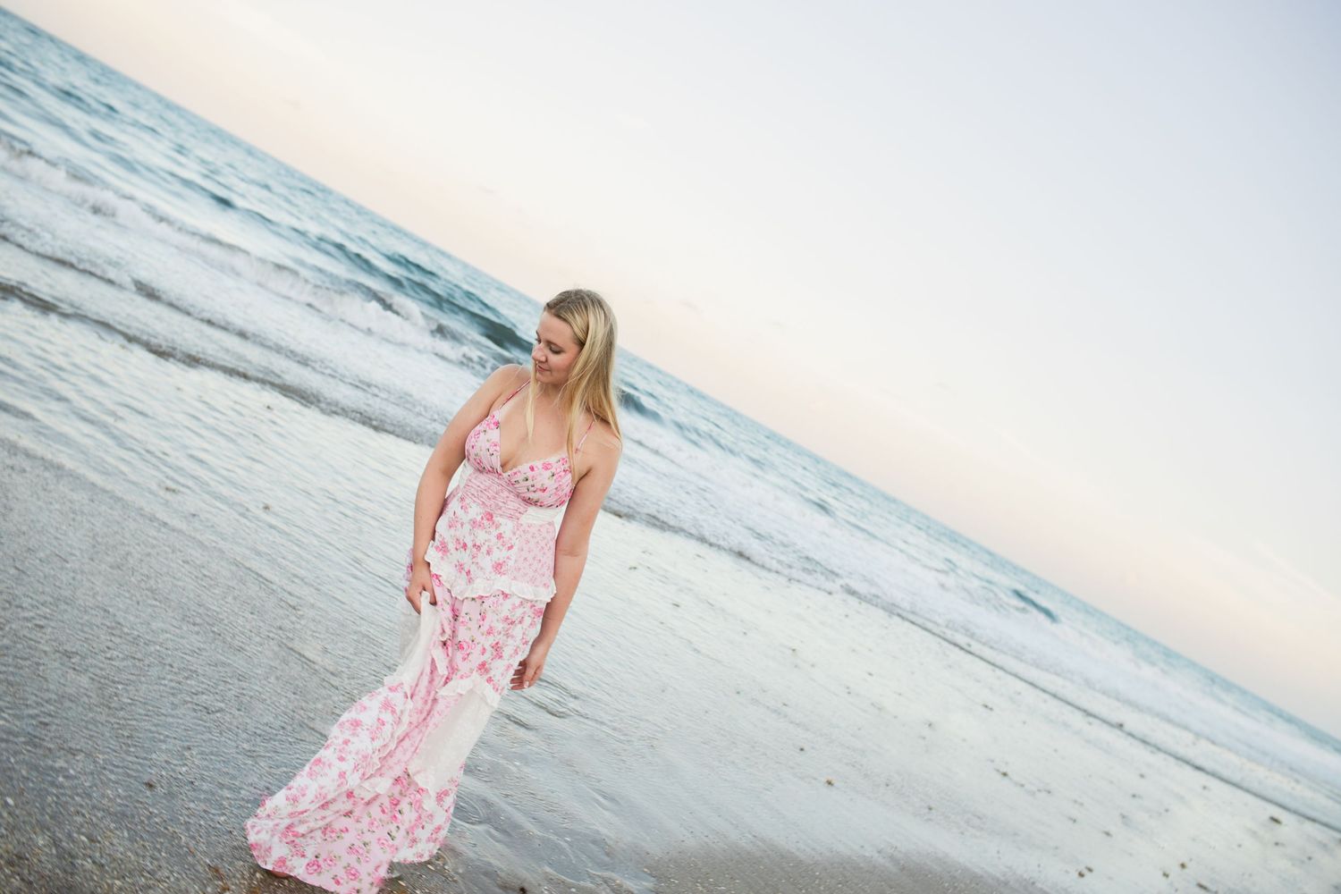 Blonde woman in pink floral maxi dress walking along sandy beach shore at sunset with ocean waves.