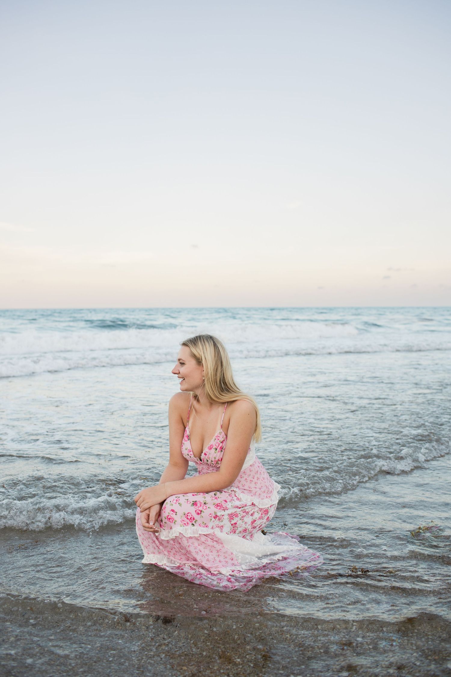 Laughing blonde woman in pink floral dress crouching at ocean's edge on sandy beach.