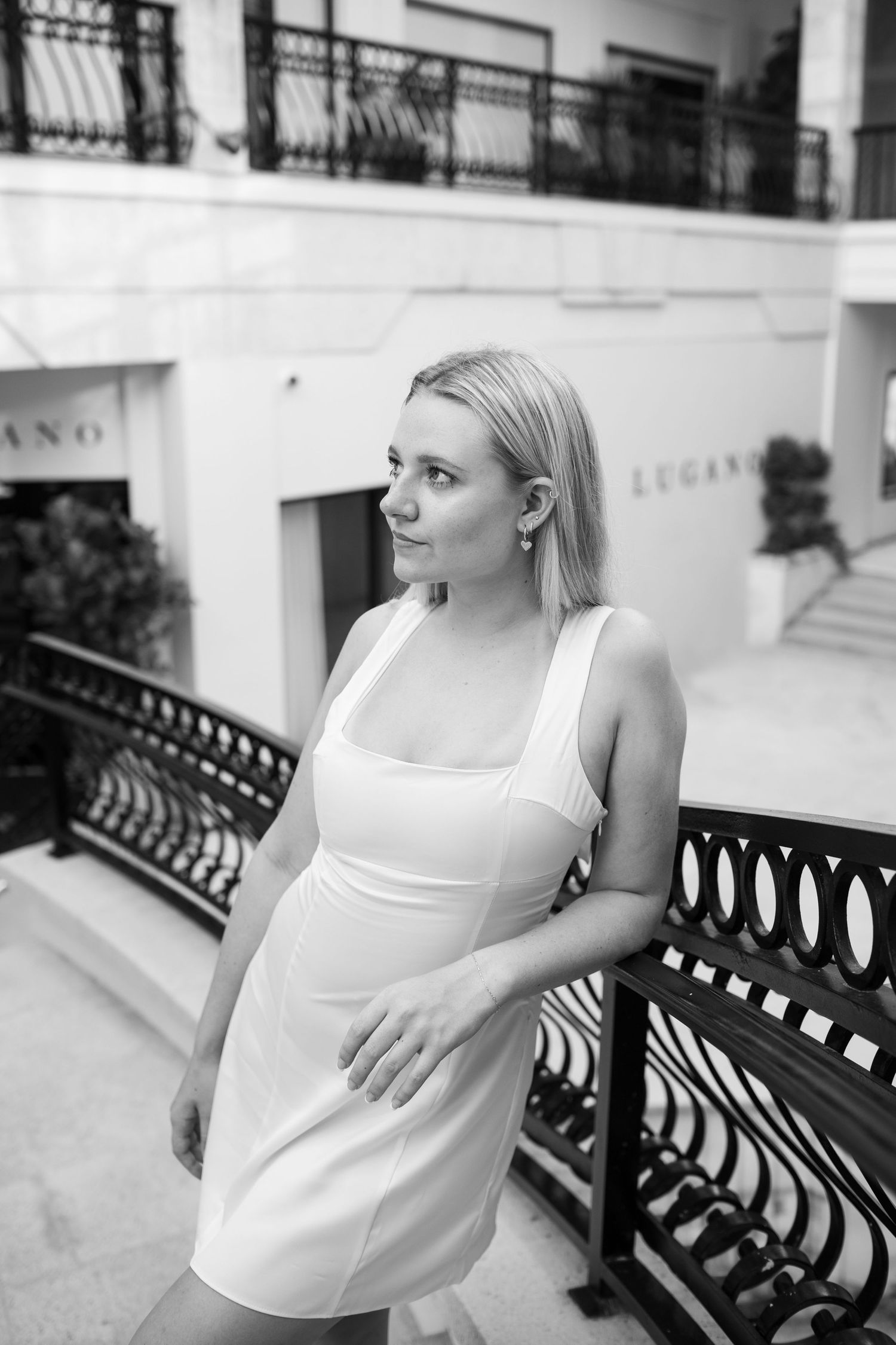 Black and white portrait of blonde woman in white dress leaning against ornate railing outside Lugano.