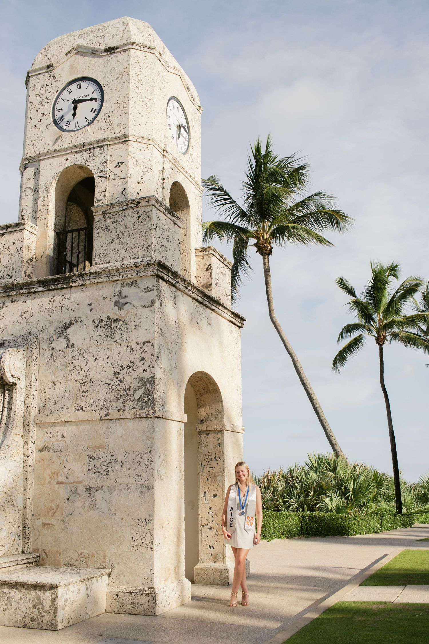 Blonde woman in white graduation dress with medal walking near historic stone clock tower with palm trees.