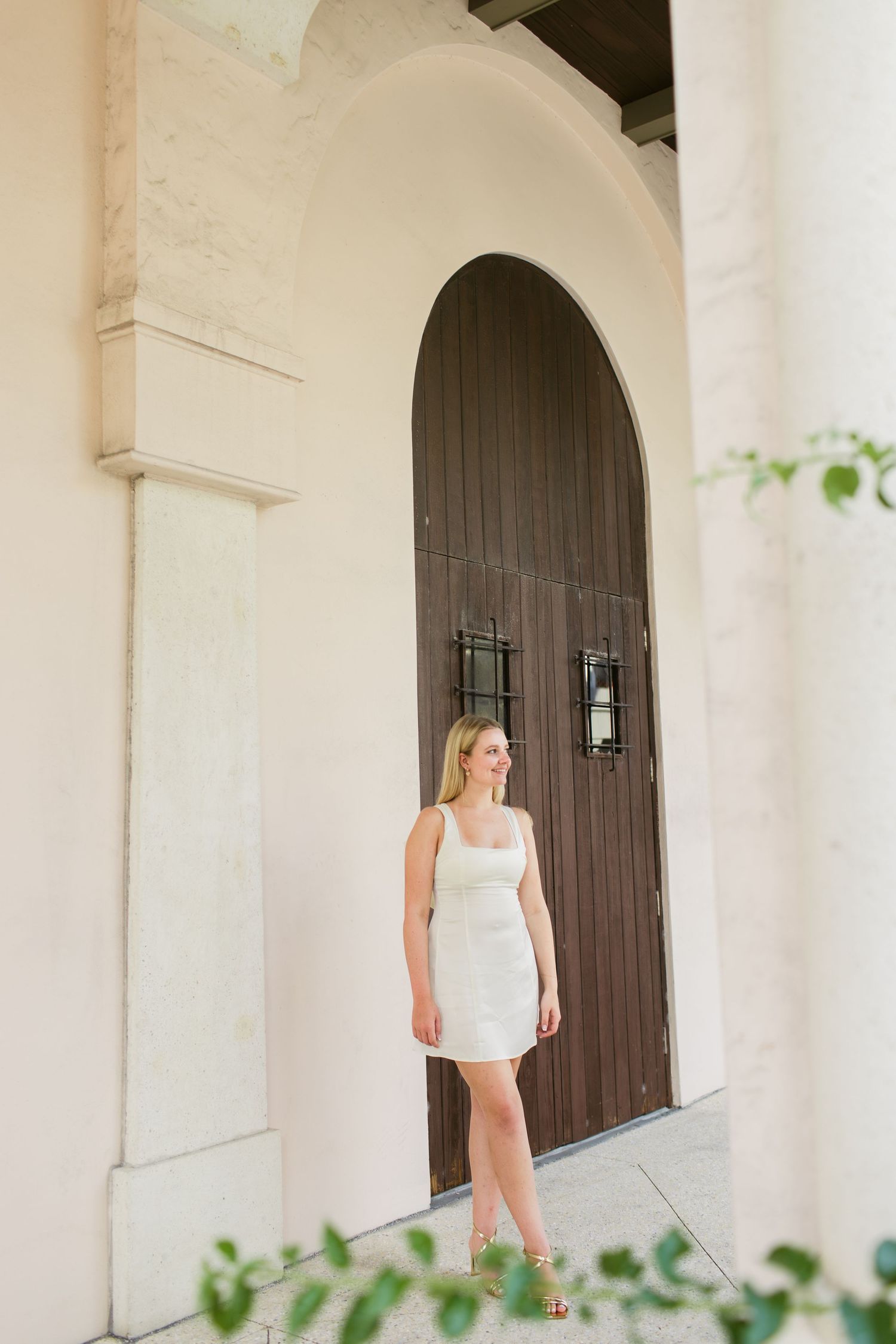 Blonde woman in white mini dress standing before arched wooden door of elegant building.