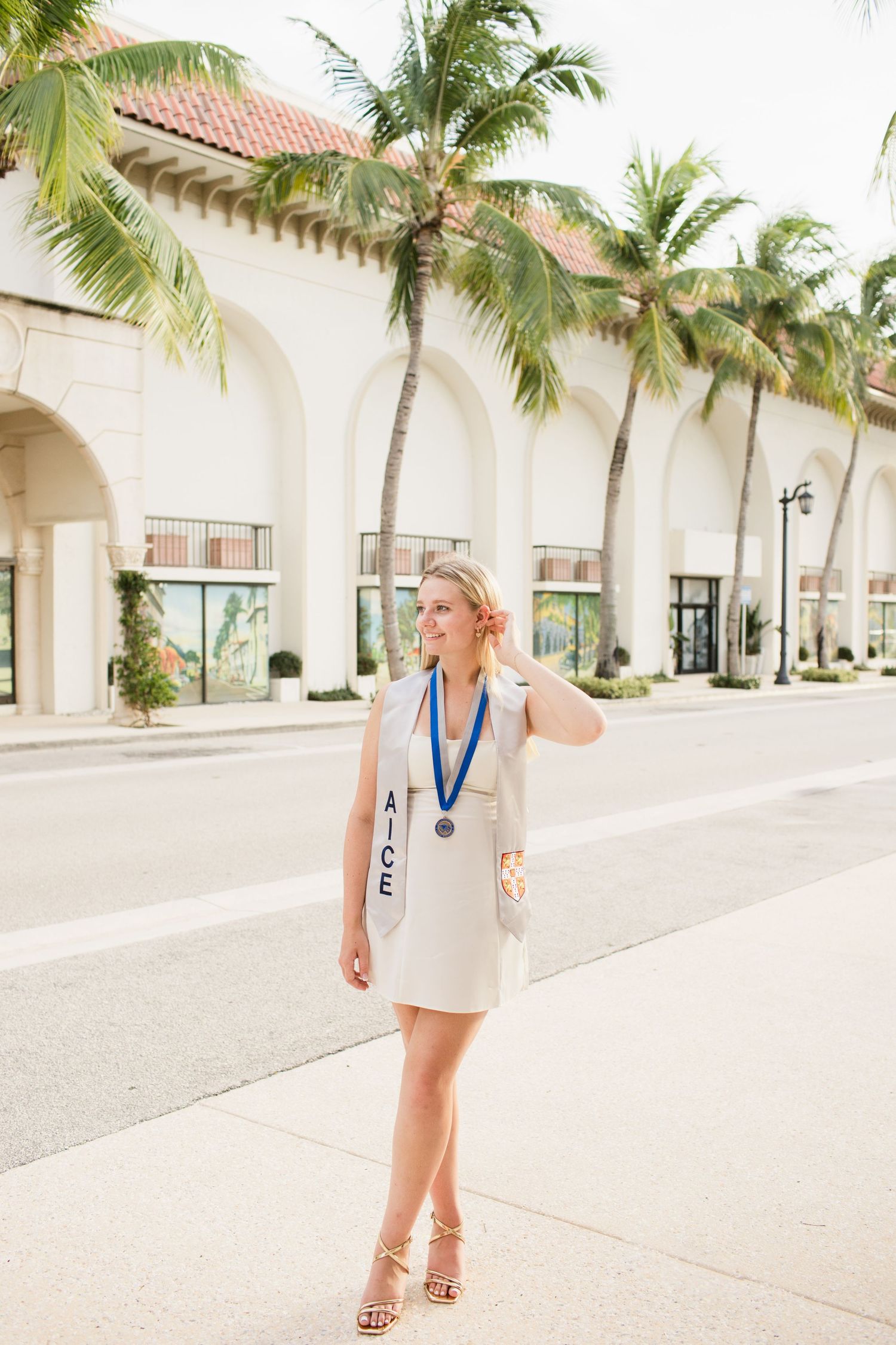 Blonde graduate in white dress with AICE sash and medal posing on a palm-lined street.