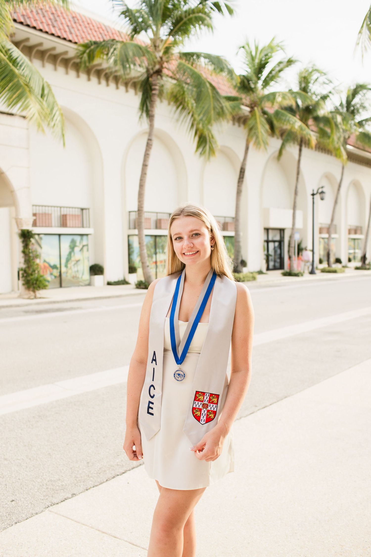 Smiling blonde graduate in white dress with AICE sash and blue medal standing on palm-lined street.