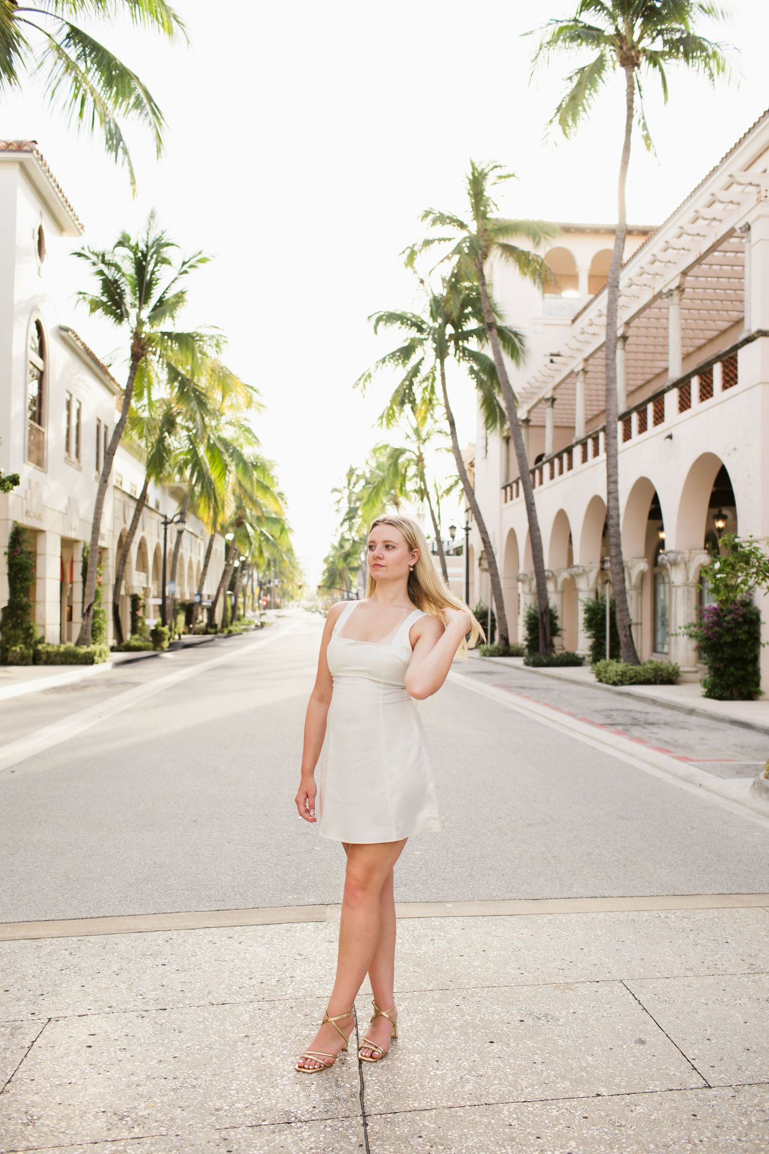 Blonde woman in white mini dress standing on palm tree-lined street of upscale tropical shopping district.
