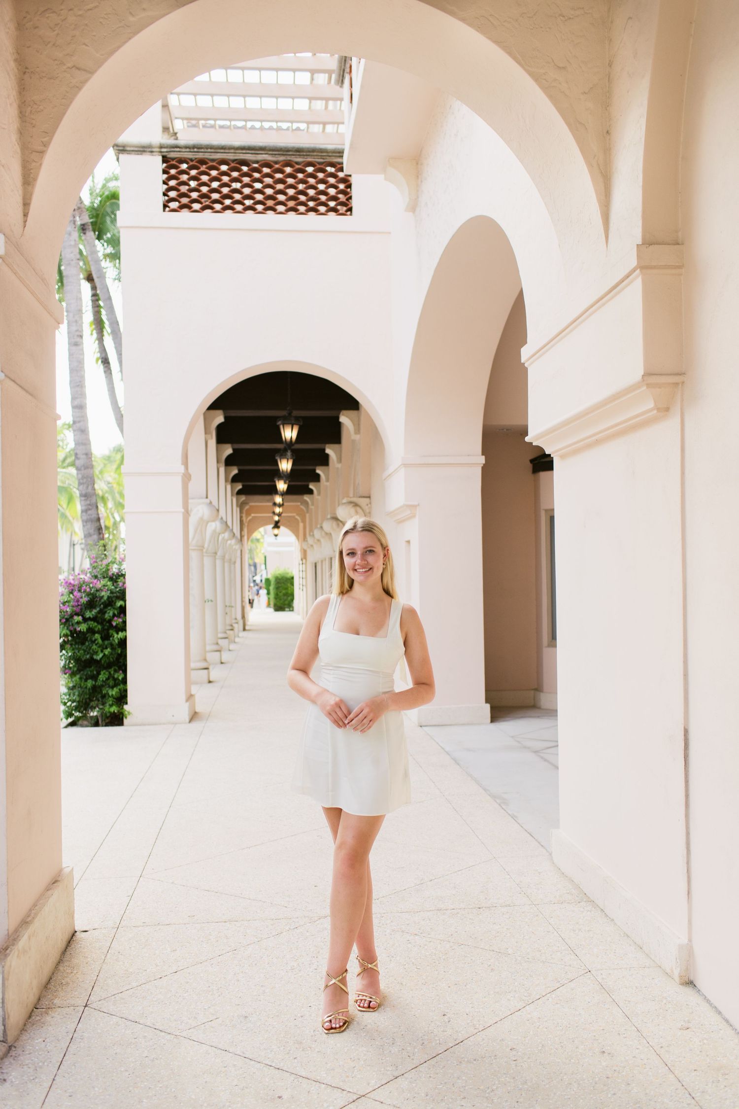Blonde woman in white mini dress posing under elegant arched colonnade of a luxury resort building.