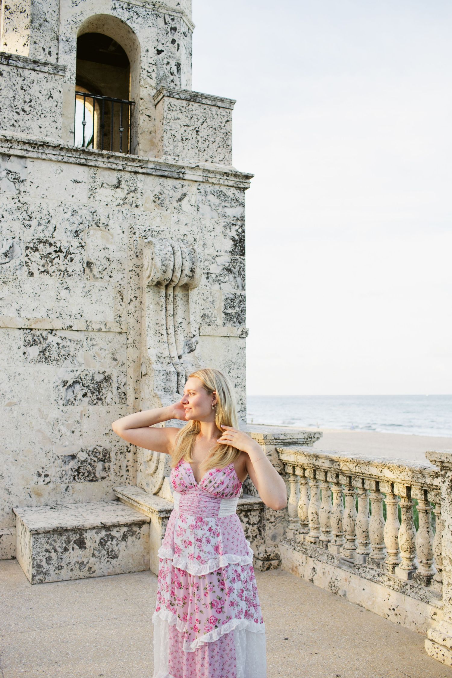 Blonde woman in pink floral maxi dress posing by weathered stone tower with ocean view.