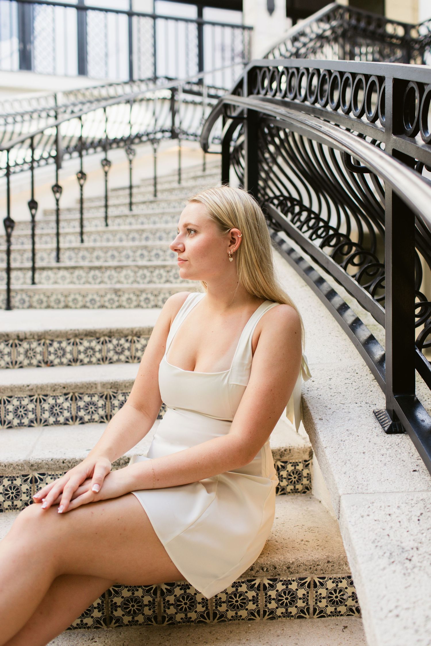 Blonde woman in cream mini dress sitting on ornate tiled staircase with decorative black iron railing.