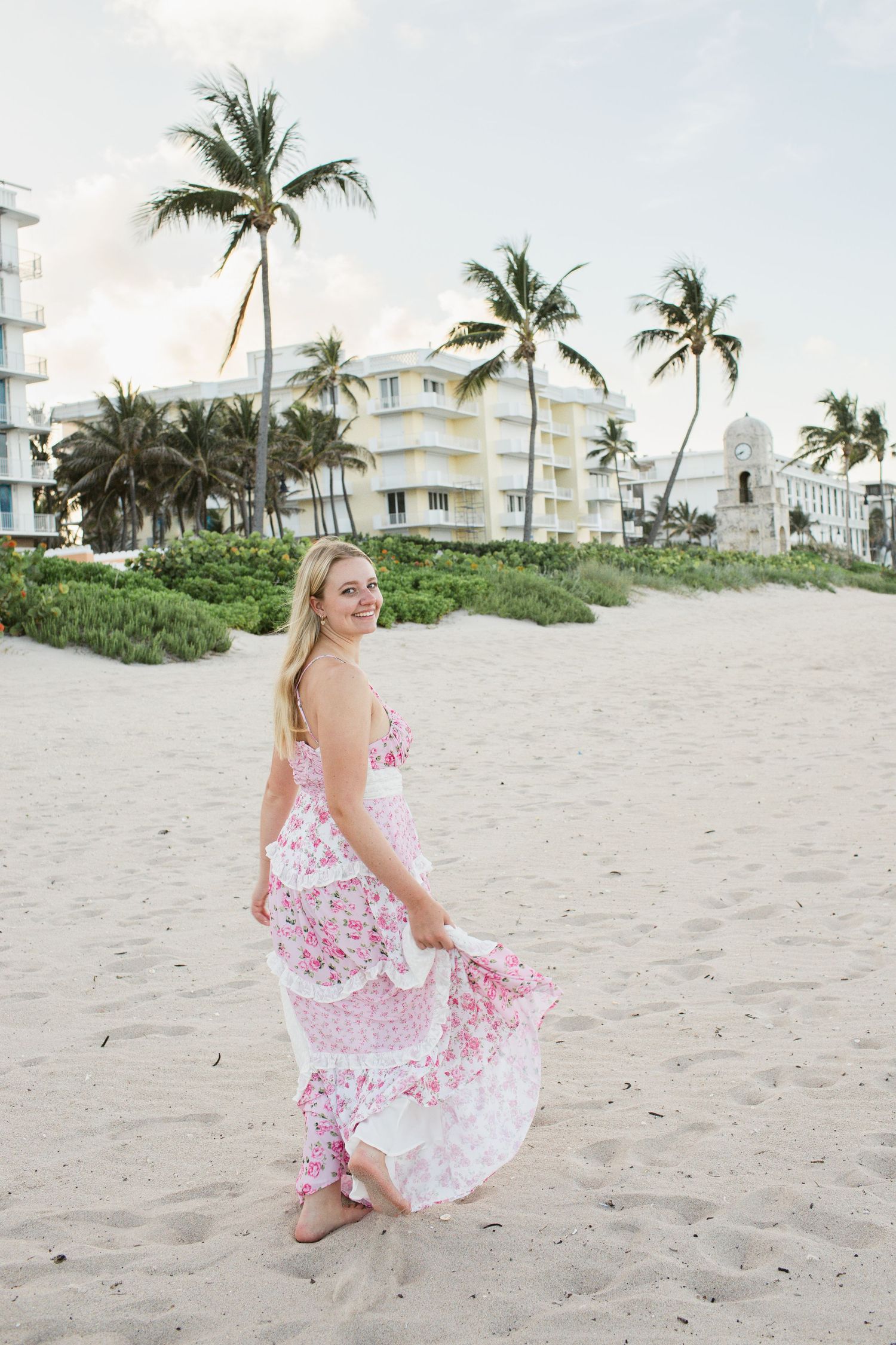 Blonde woman in pink floral maxi dress twirling on sandy beach with palm trees behind.