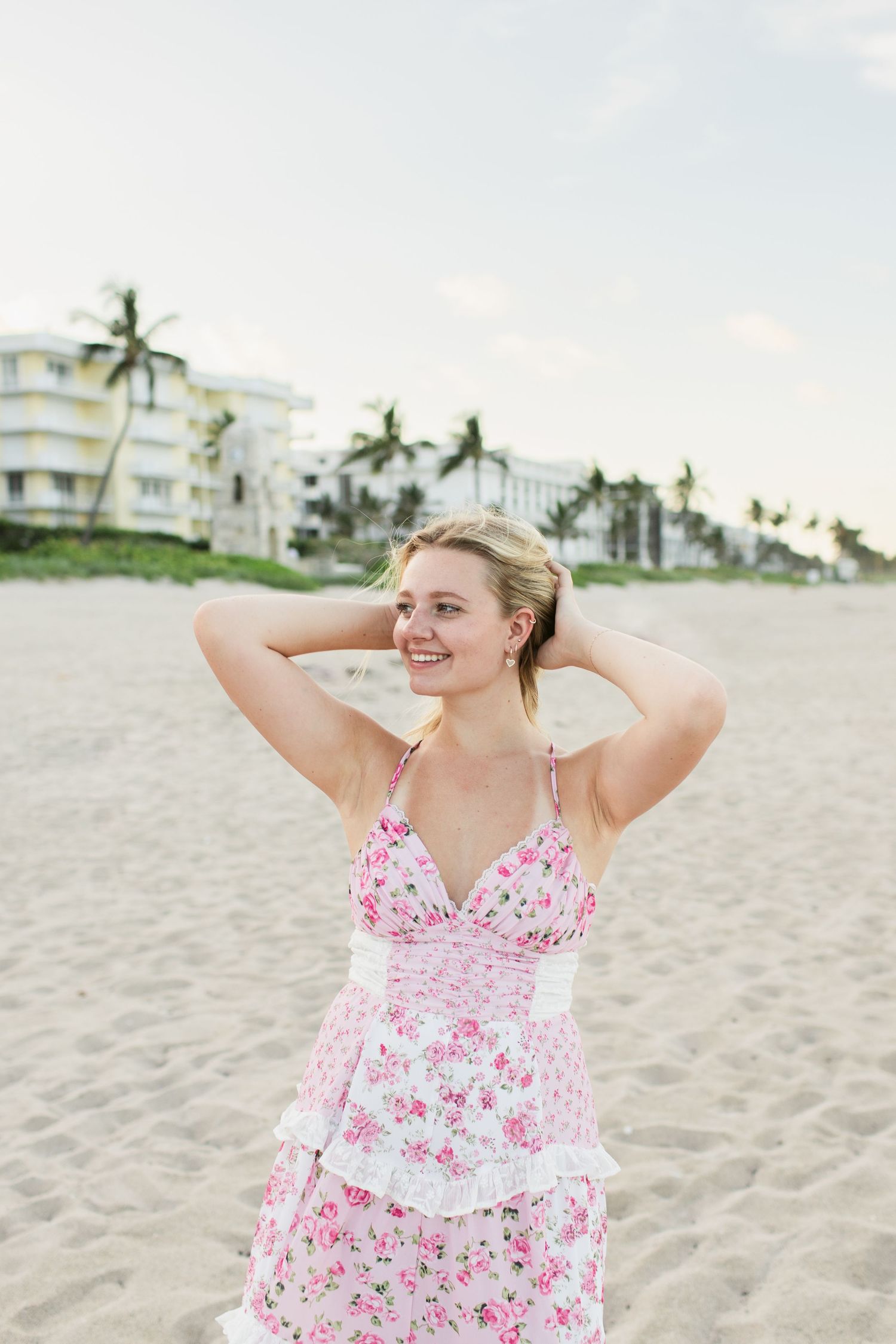 Smiling blonde woman in pink floral dress with hands in hair on sandy beach.