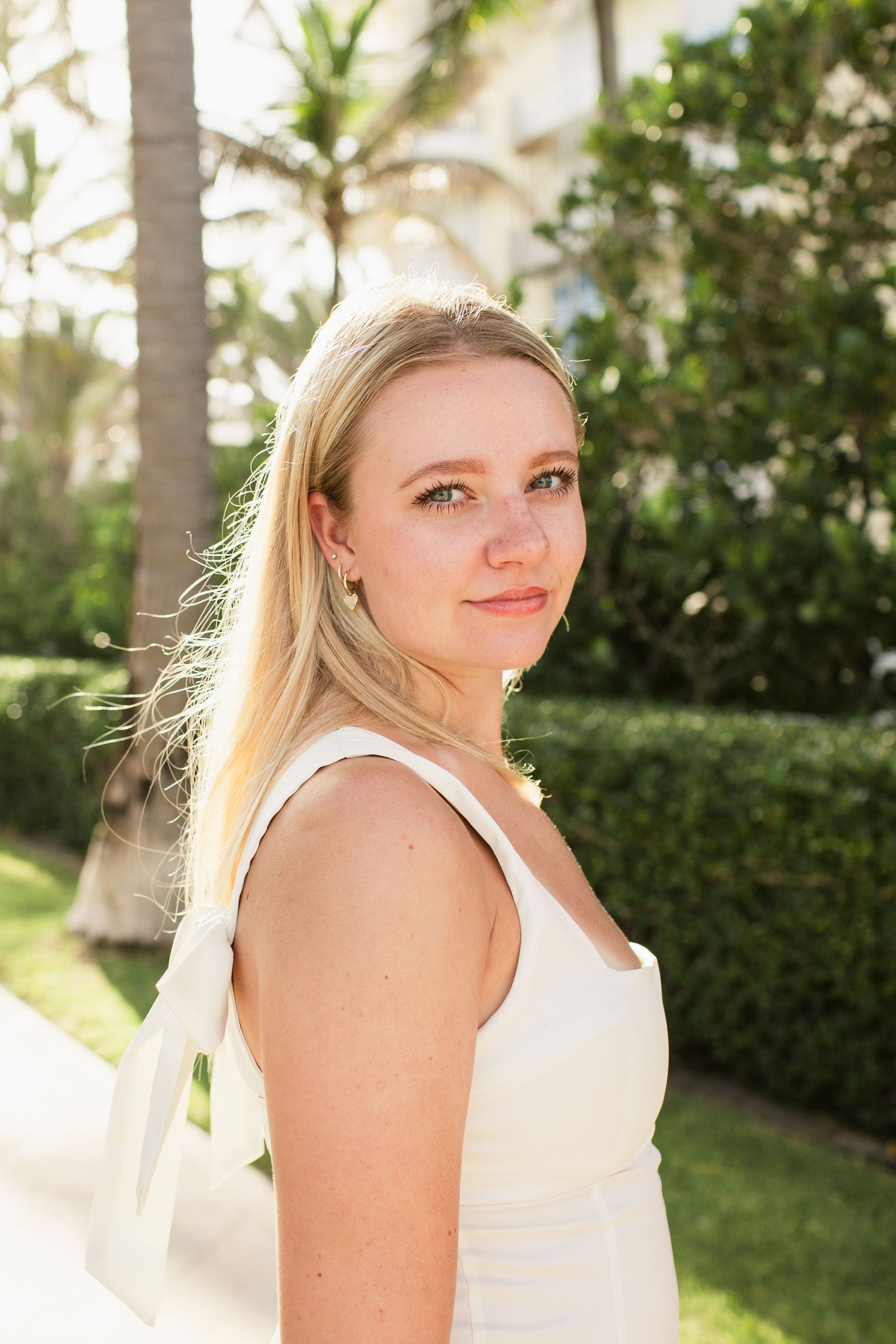 Young blonde woman in white sundress smiling outdoors with palm trees and green hedges behind her.