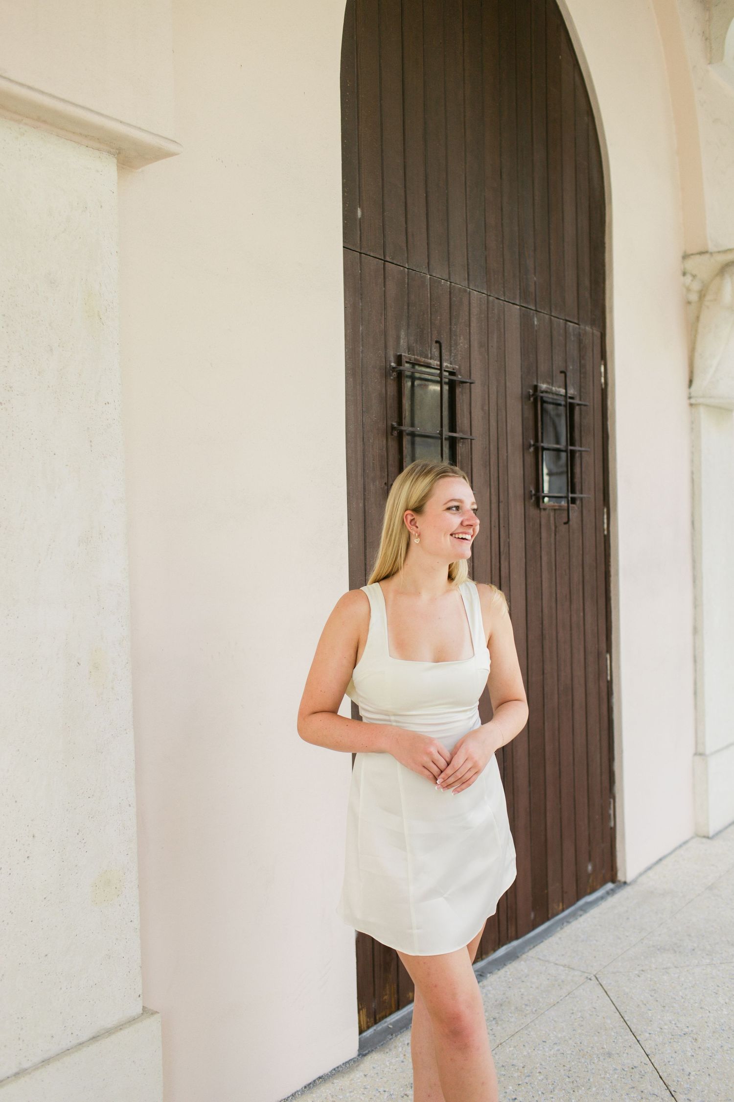 Smiling blonde woman in white mini dress leaning against wall near arched wooden door.
