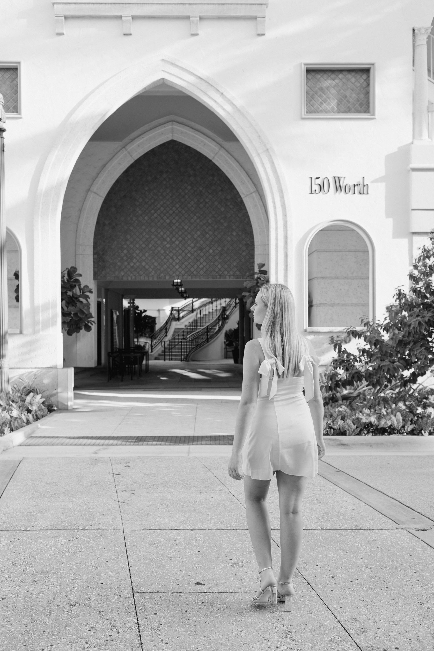 Blonde woman in white romper standing before arched entrance of 150 Worth Avenue building in black and white photo.