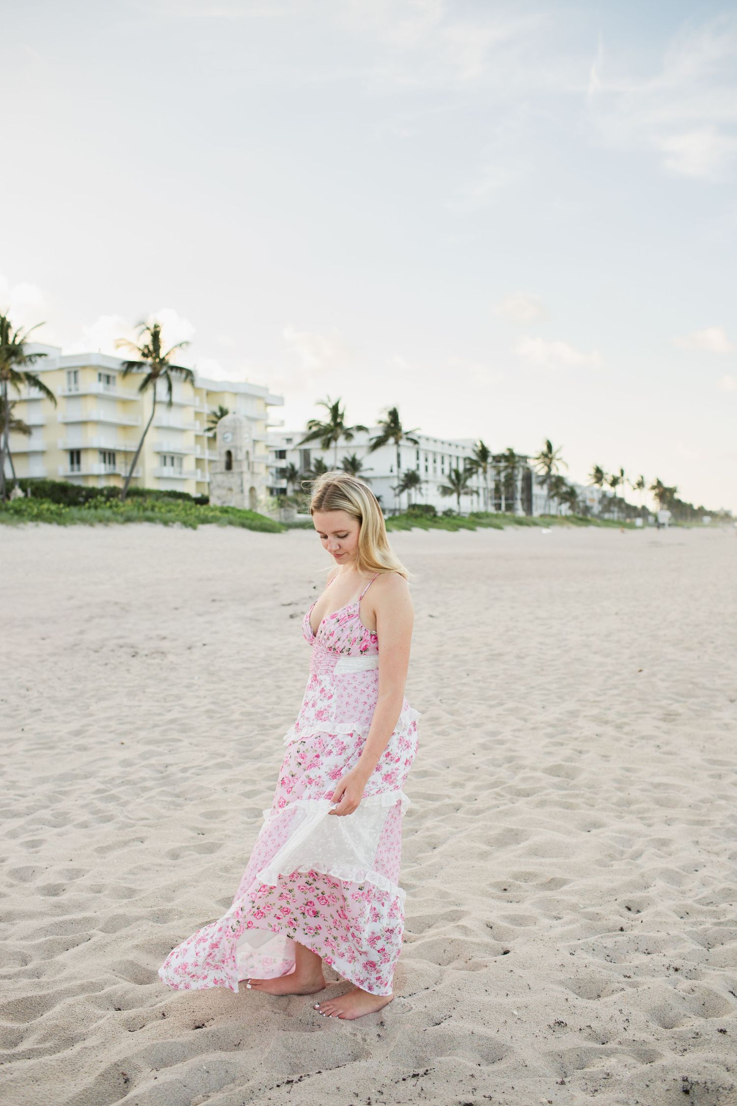 Blonde woman in pink floral maxi dress walking barefoot on sandy beach at dusk.