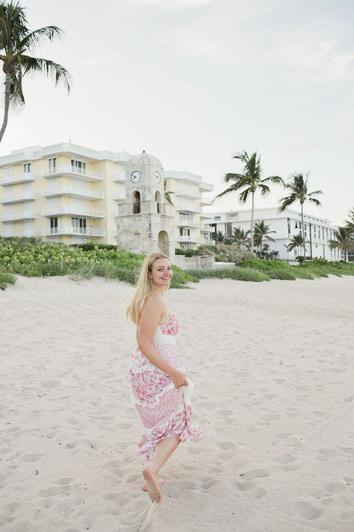 Smiling blonde woman in pink floral dress walking barefoot on sandy beach near buildings.