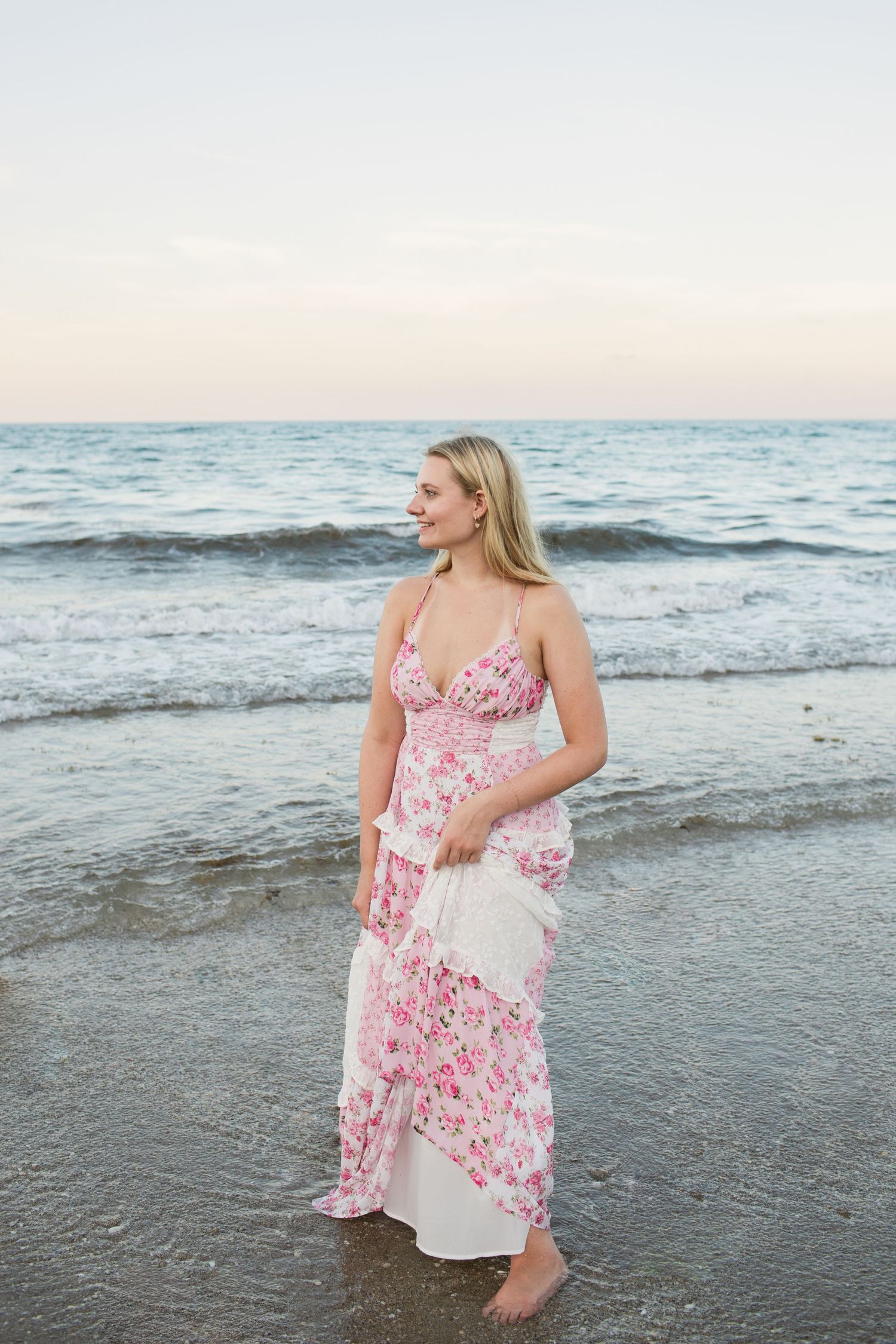 Smiling blonde woman in pink floral maxi dress standing barefoot at the ocean shoreline at dusk.