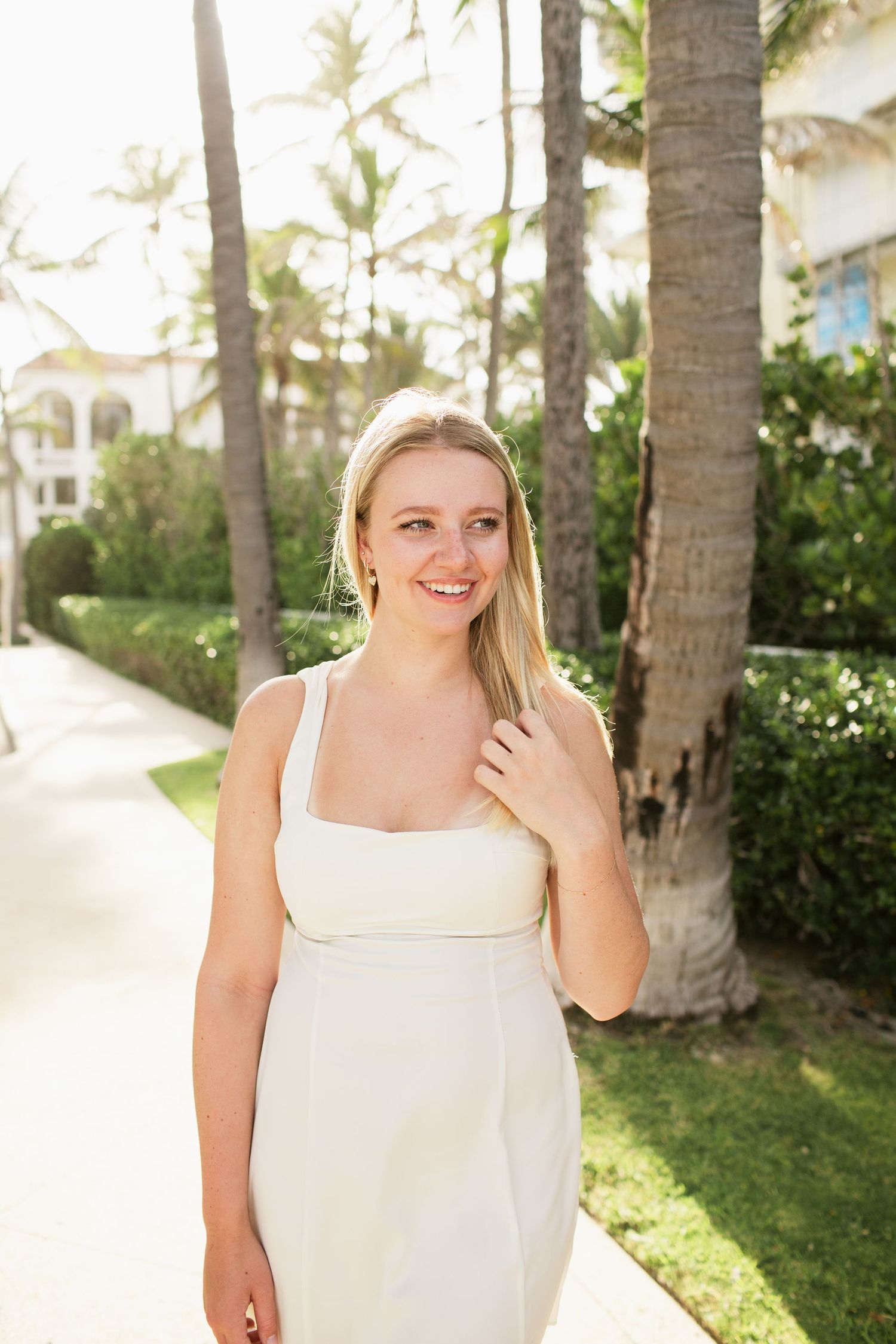 Smiling blonde woman in white sundress walking along palm tree-lined tropical resort pathway.