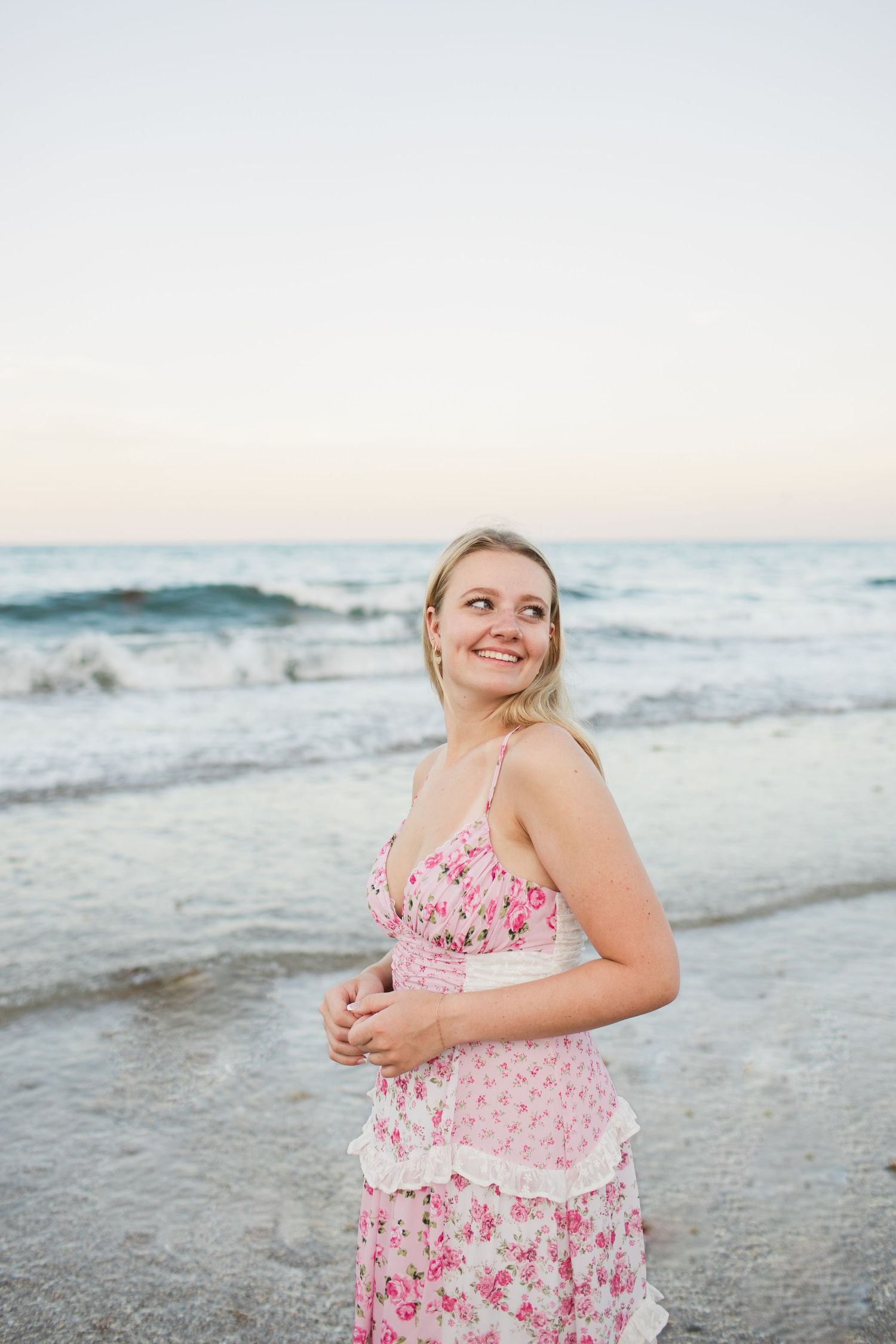 Joyful blonde woman in pink floral dress laughing on beach at dusk by ocean.
