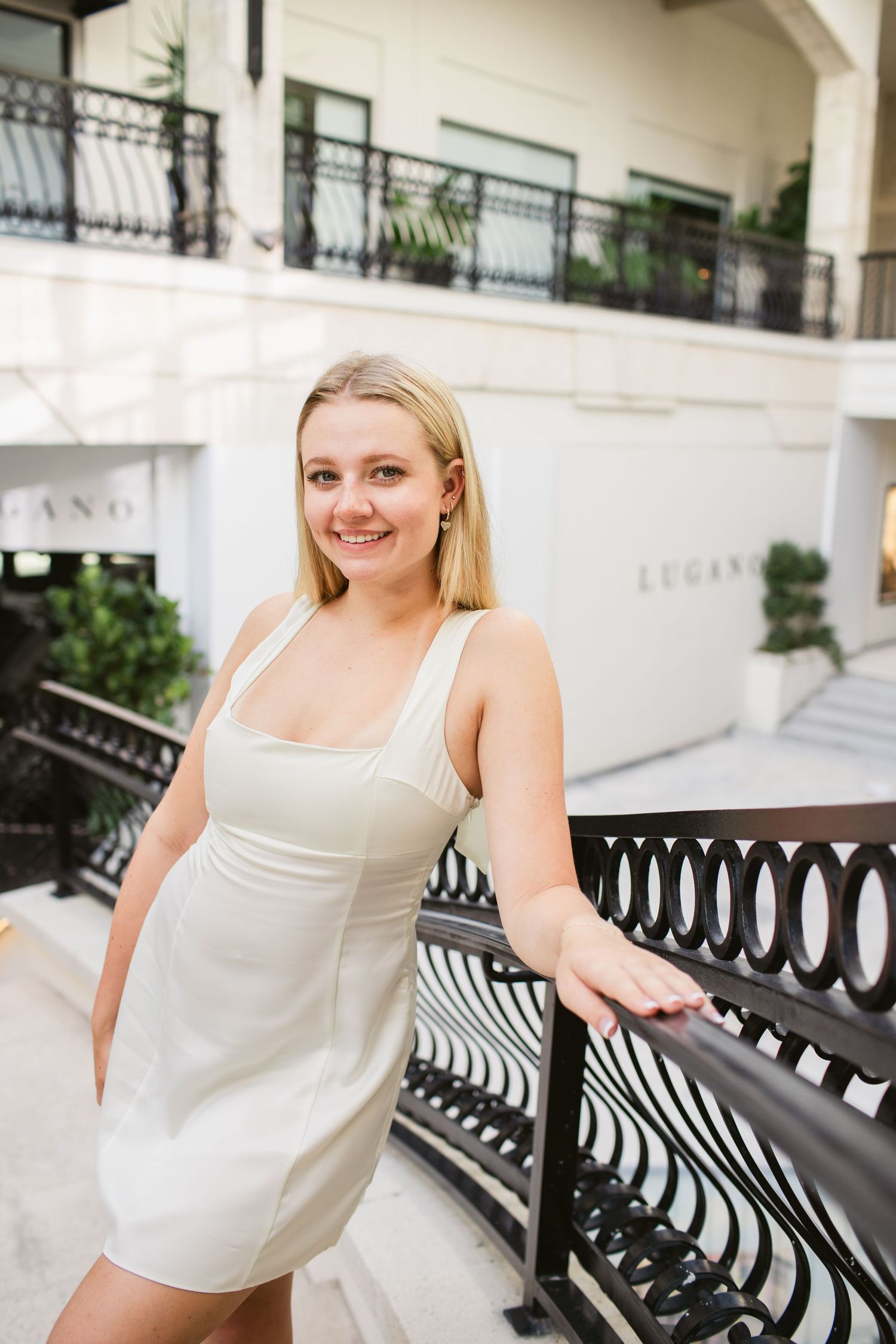 Smiling blonde woman in white dress leaning on black iron railing outside Lugano luxury boutique.