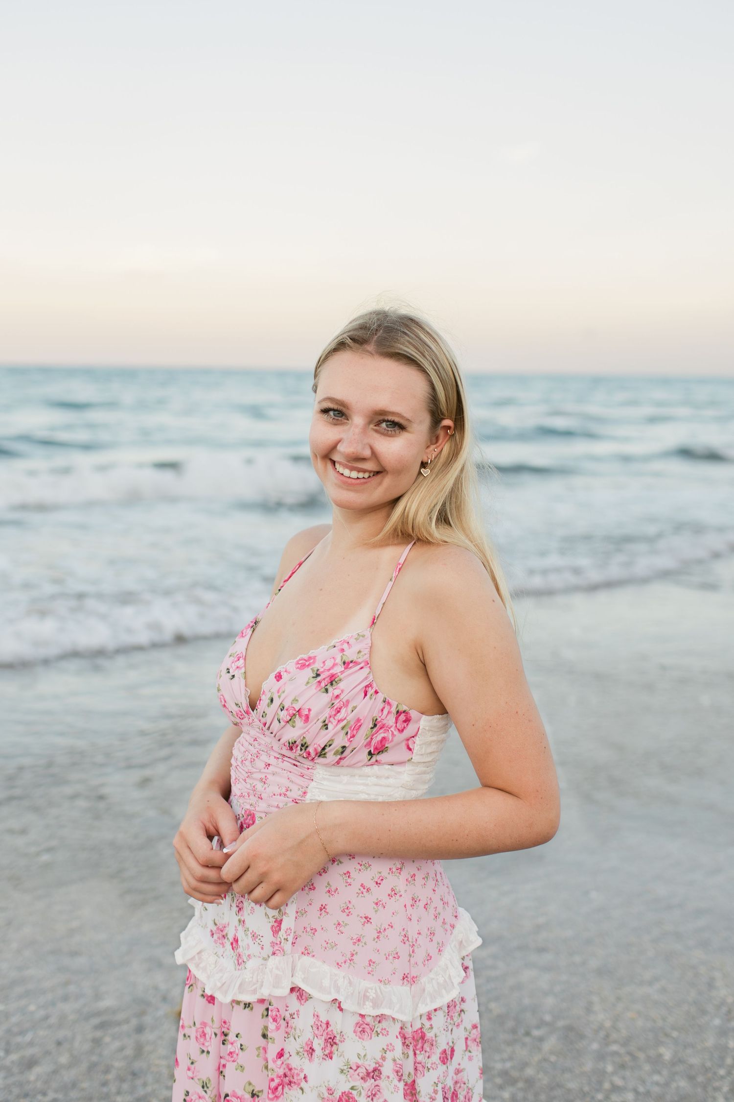 Smiling blonde woman in pink floral sundress standing on beach with ocean waves behind her.