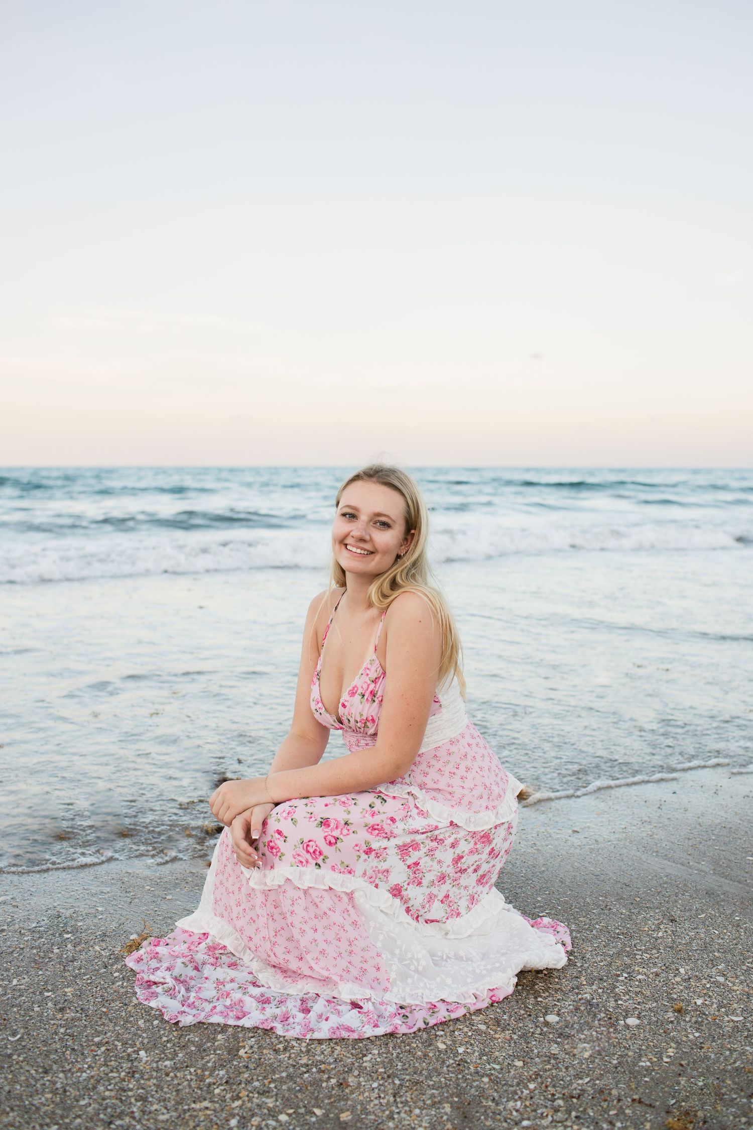 Smiling blonde woman in pink floral maxi dress sitting on wet sand at beach.