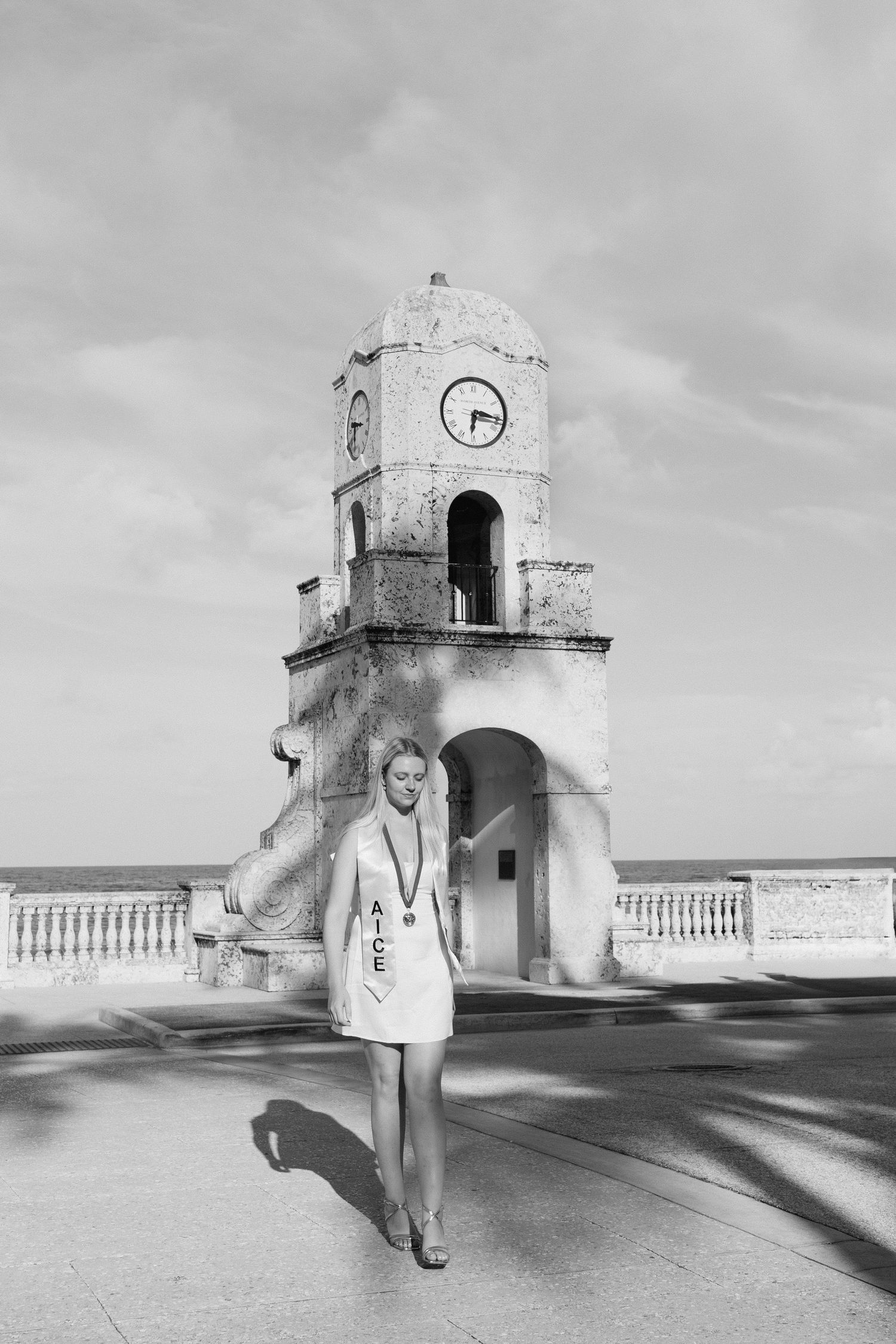Black and white photo of young woman with graduation medal standing before historic clock tower by the sea.