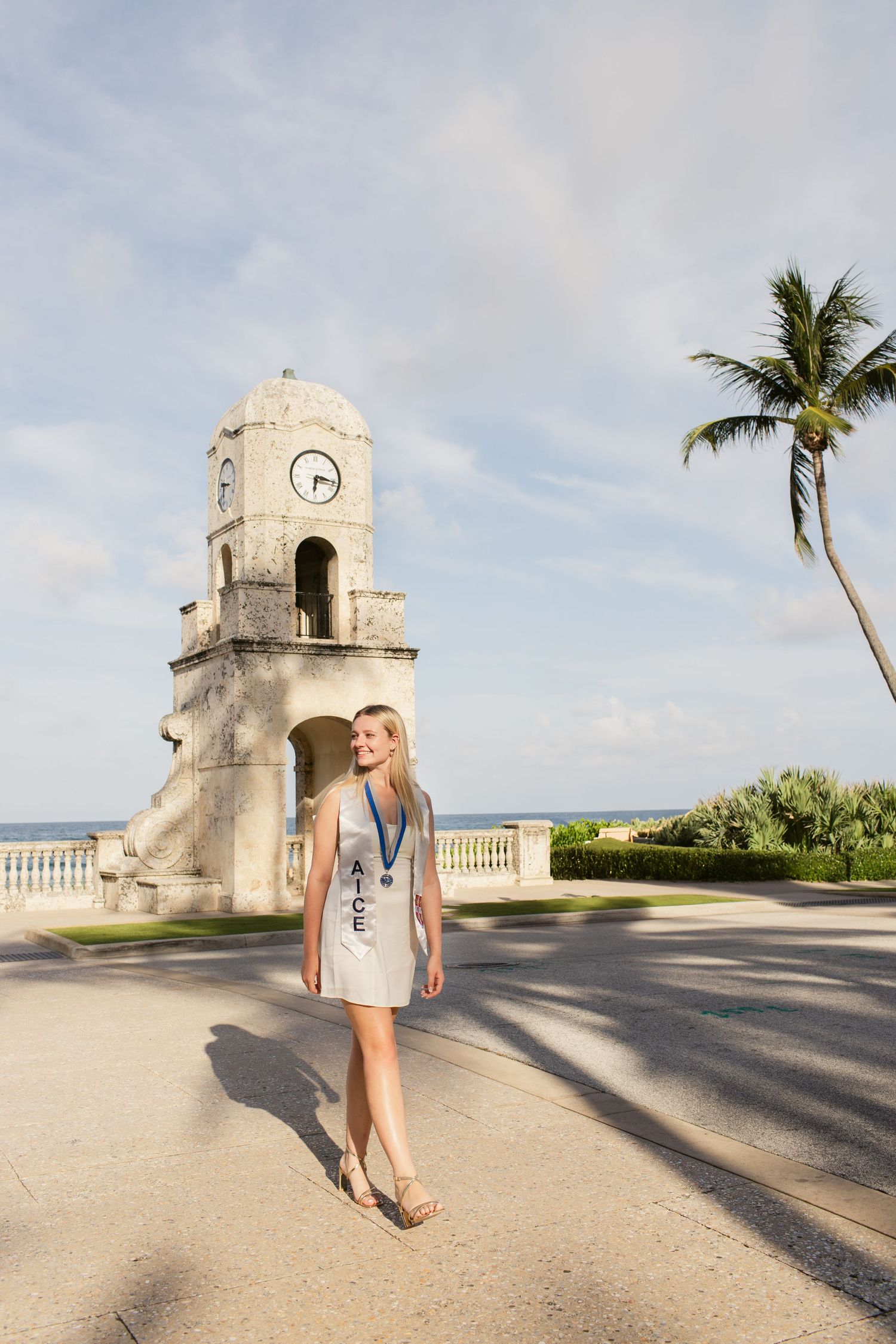 Young blonde woman in white dress with AICE sash and medal standing near historic clock tower with palm trees.