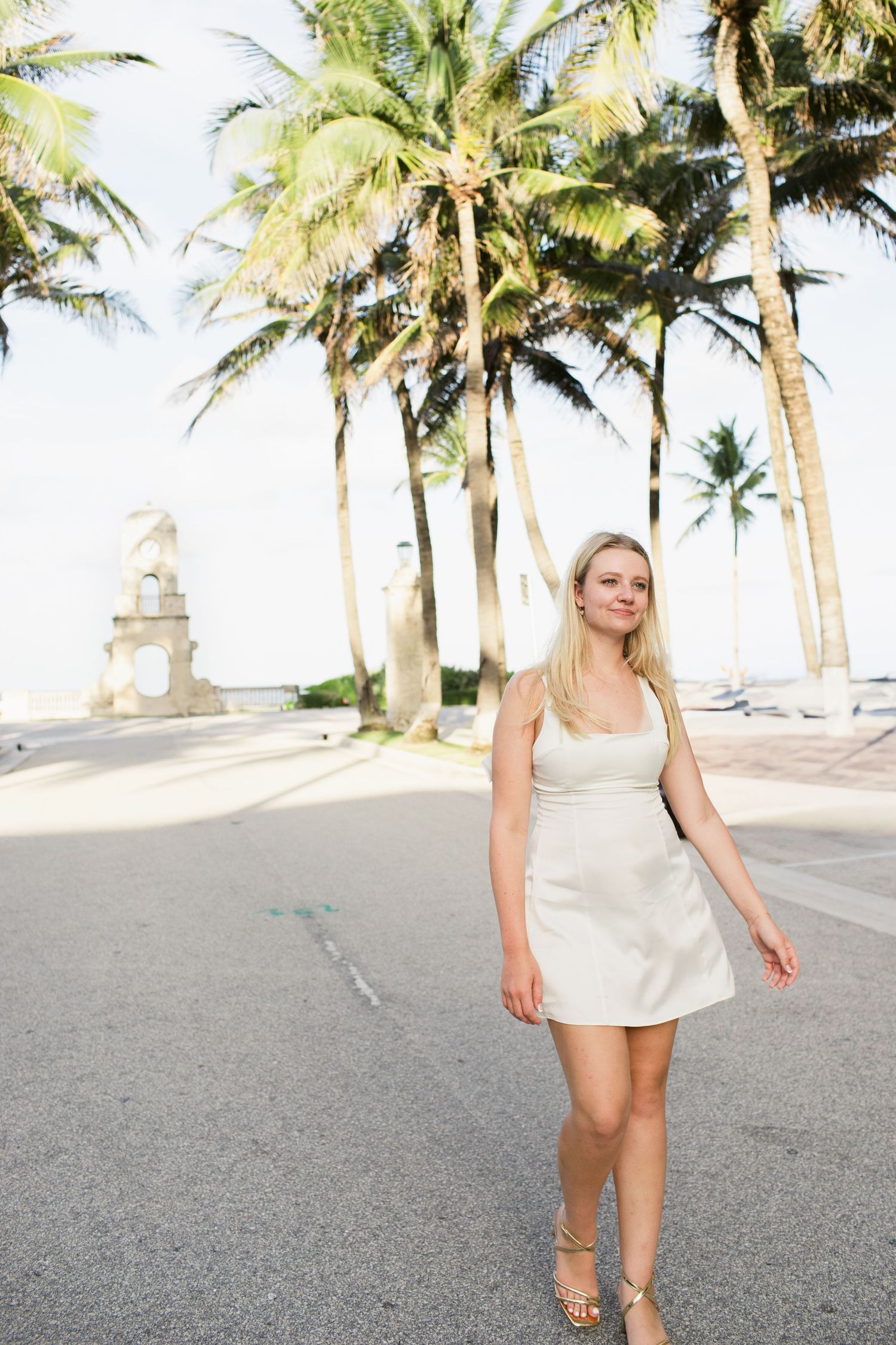 Blonde woman in a white mini dress walking along a palm-lined street near a clock tower.