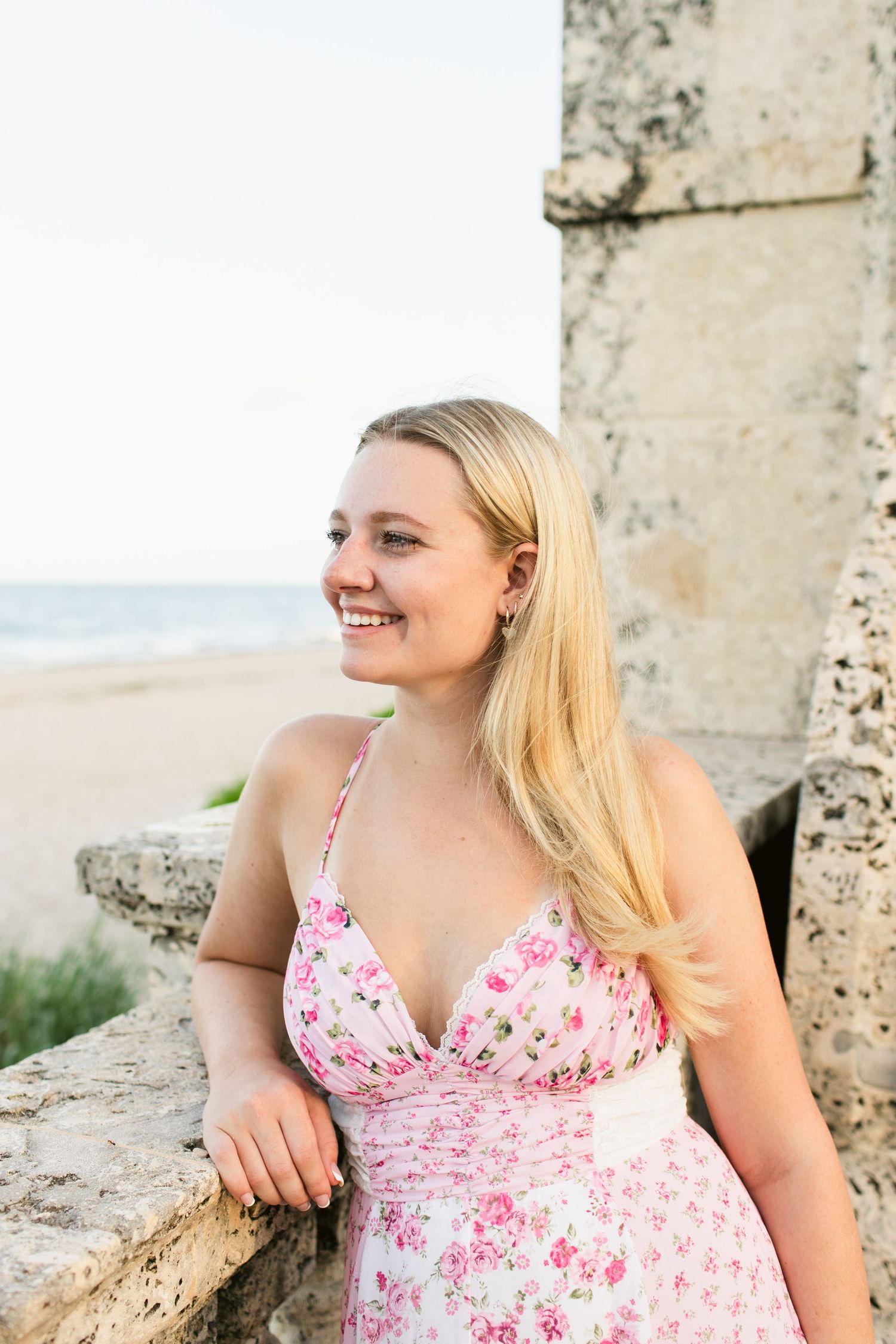 Smiling blonde woman in pink floral halter dress leaning on stone balustrade by beach.