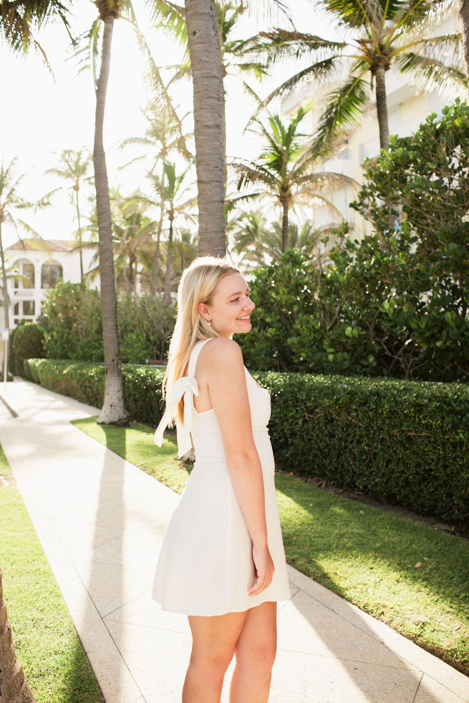 Smiling blonde woman in white mini dress standing on sunny tropical walkway lined with palm trees.