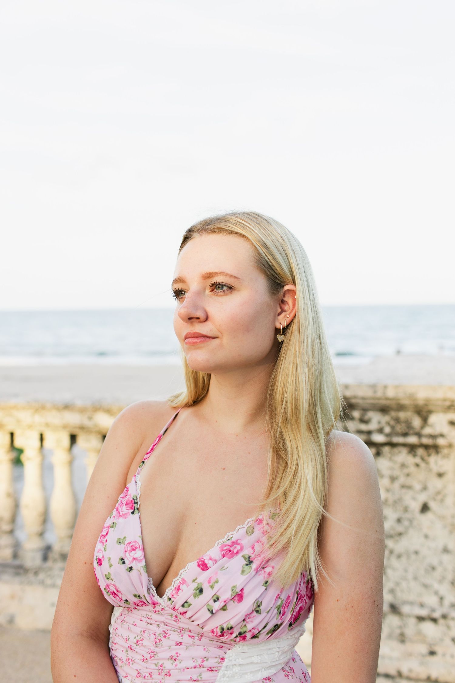 Blonde woman in pink floral halter dress gazing thoughtfully with ocean in background.